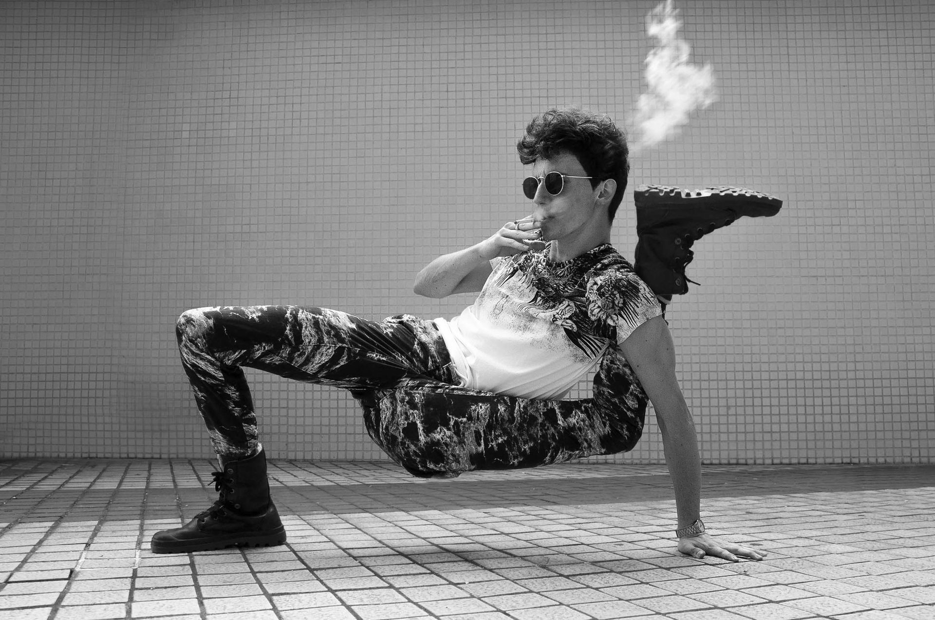 Man in patterned clothing doing a dance move, smoking. Black and white photo with a brick wall backdrop.