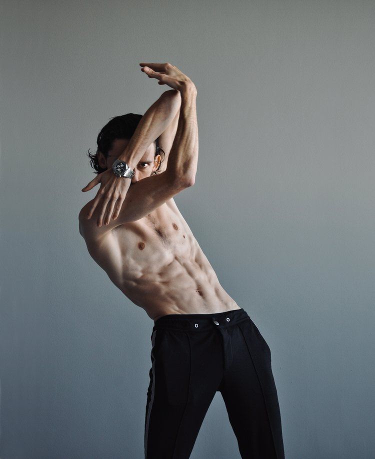 Shirtless person in black pants, arms overhead, angled pose. Gray backdrop.