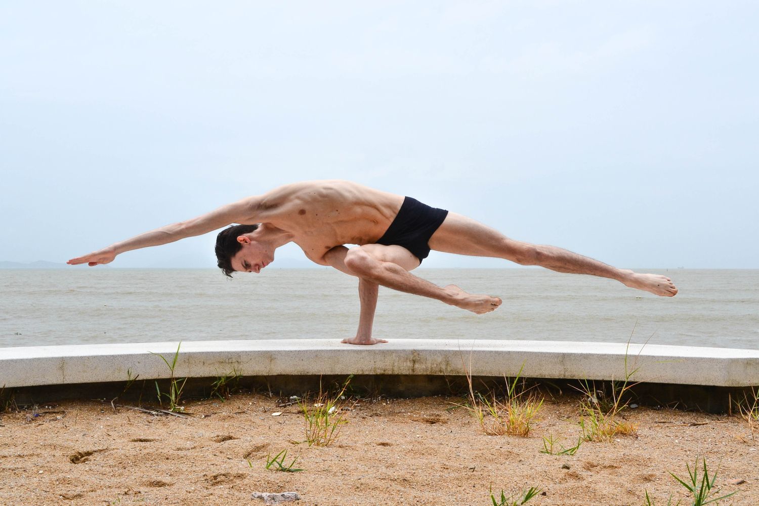 Man in black shorts balances in yoga pose on a ledge, arms outstretched, near the ocean.