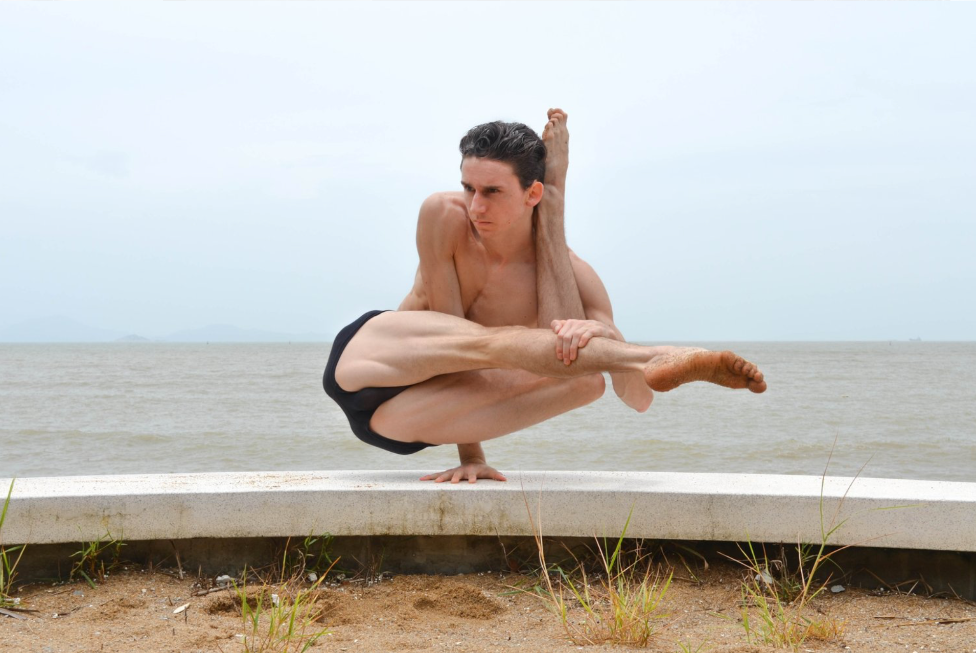 Man in yoga pose on a seaside ledge, balancing on one hand with legs crossed and bent upward.