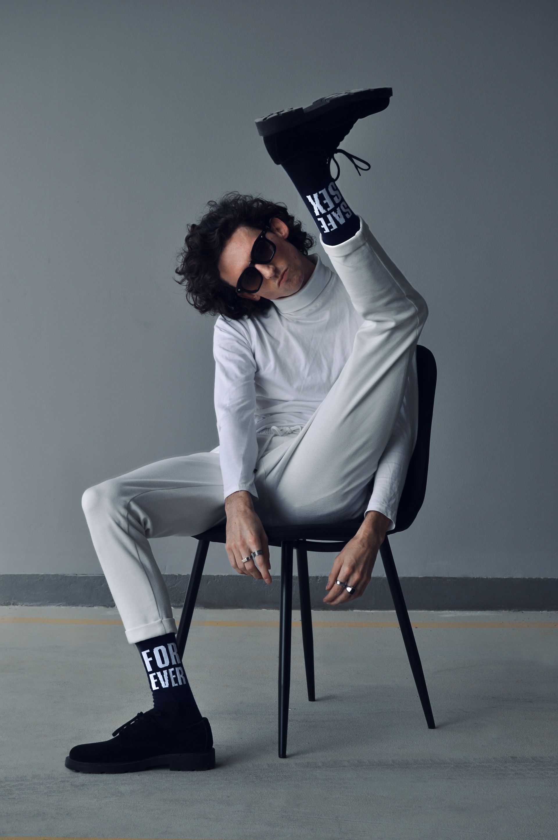 Person in white outfit, sunglasses, and black socks, leg raised, sitting on a black chair. Grey background.