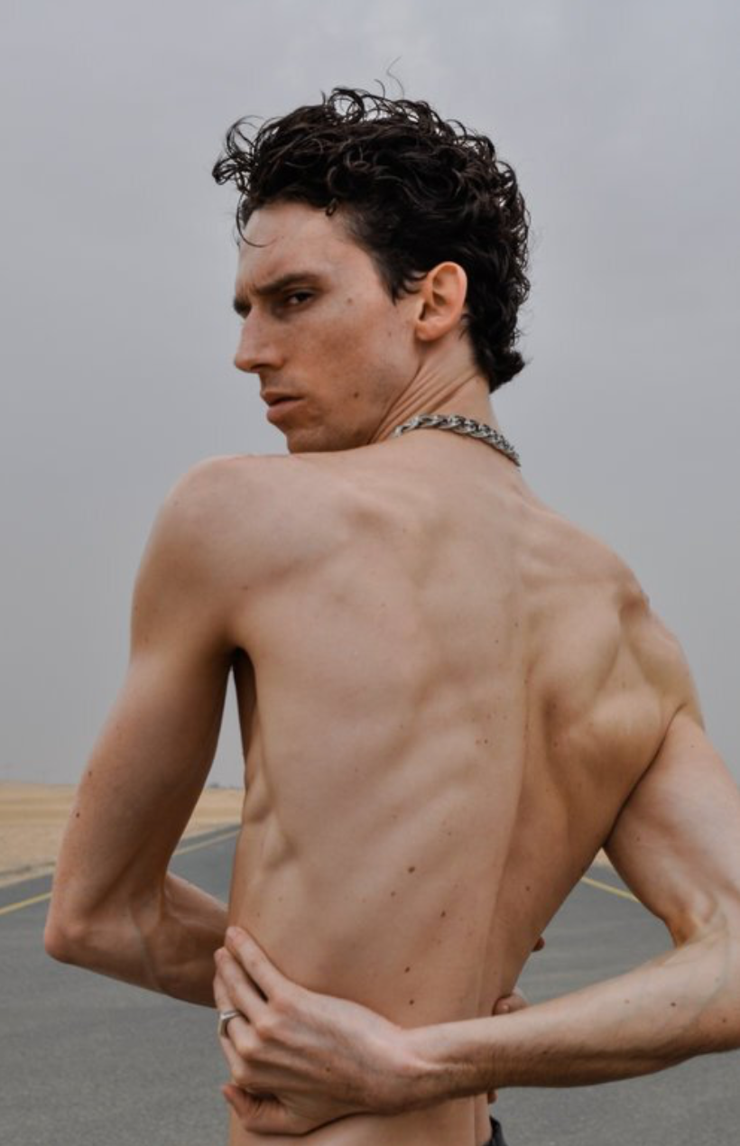 Man with curly hair, shirtless, turning to look over his shoulder; chain necklace, gray sky, open road.
