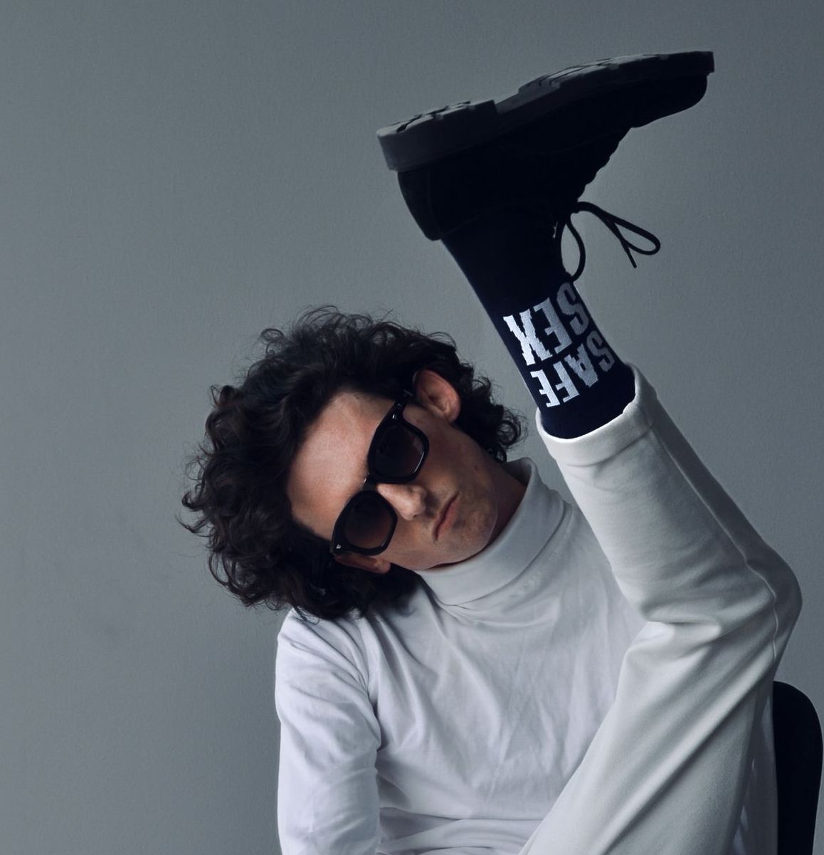 Person wearing sunglasses, turtleneck, and raising a leg, showing sock that reads 