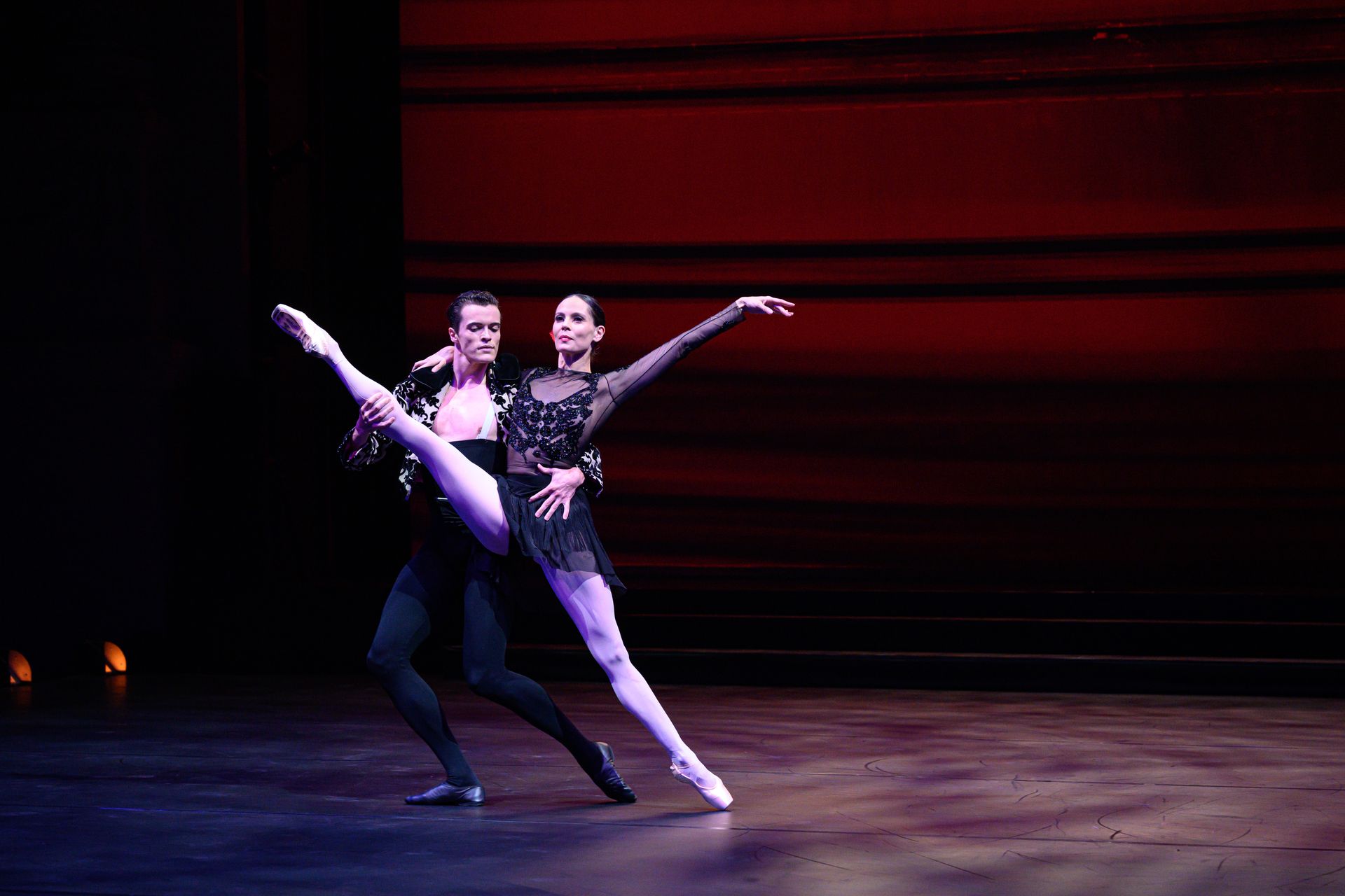 Ballet dancers on stage. Man supports woman in split. Dark setting, red background.