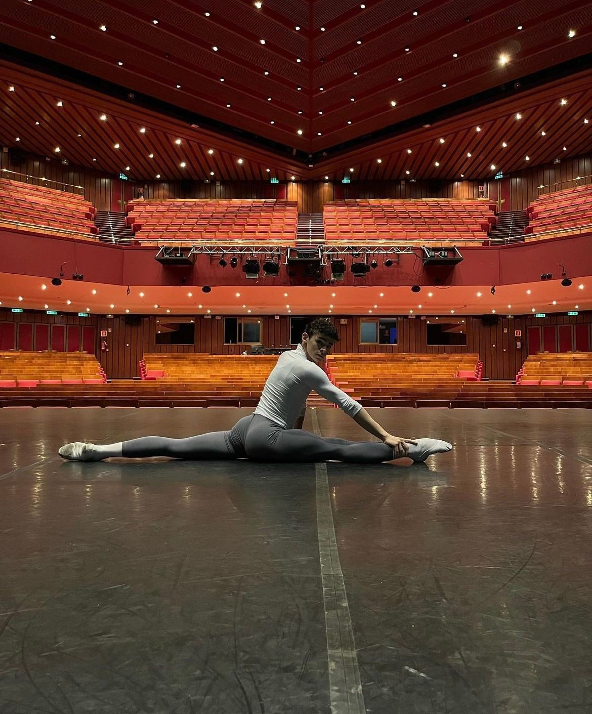 Person in a grey outfit doing the splits on a stage, reaching for their foot, in a red theater.