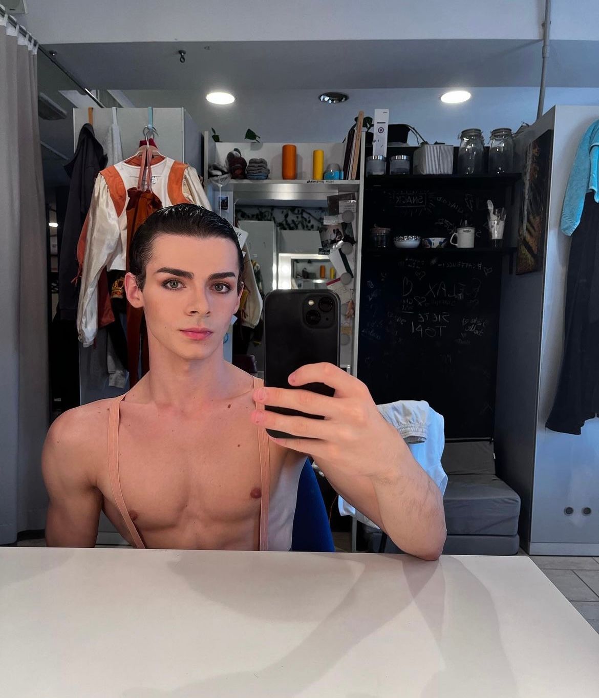 A shirtless person with makeup takes a mirror selfie in a dressing room with costumes visible.