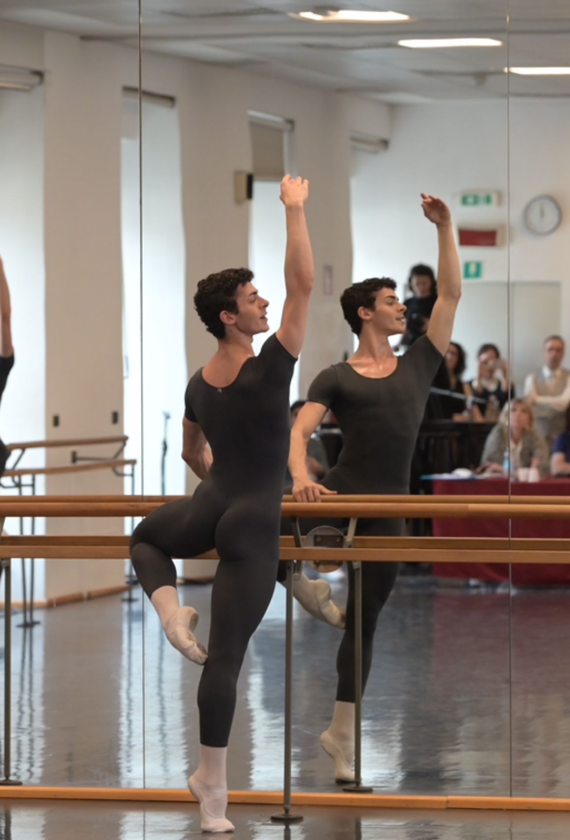 Man in dark leotard, ballet pose at barre, reflected in mirror. Indoor studio setting.