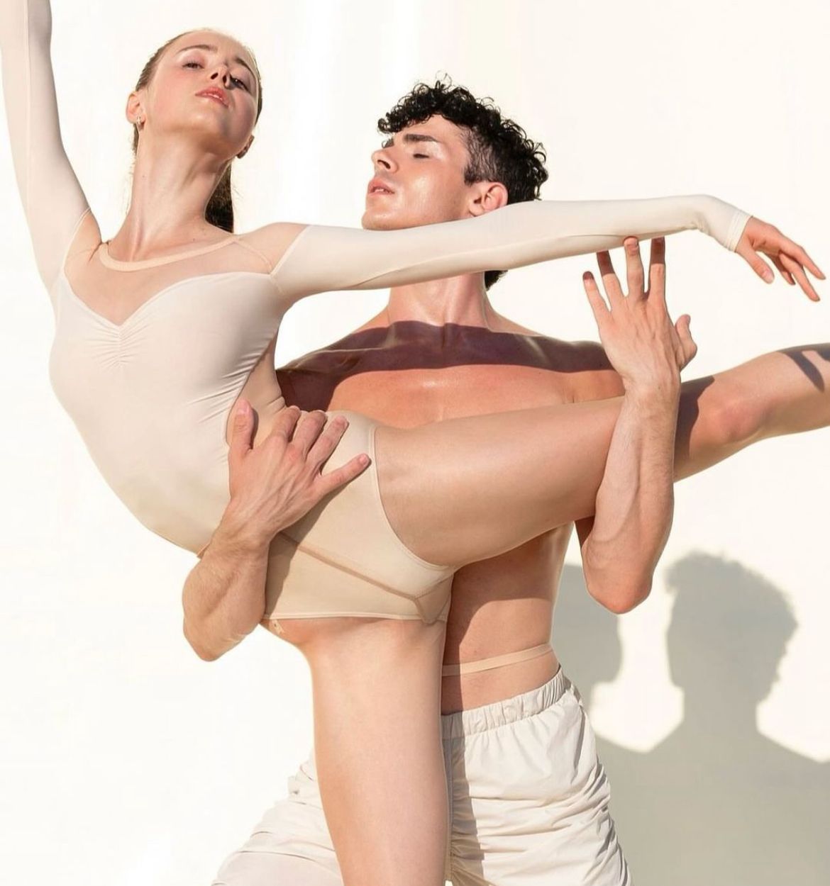 Ballet dancers in a lift: woman in beige bodysuit, man shirtless. Bright, white backdrop.