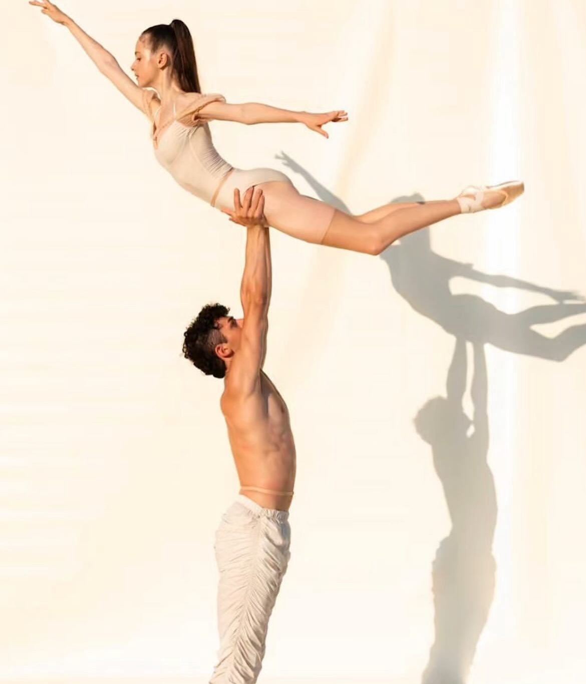 Male dancer lifts a ballerina. She's extended above him with one arm reaching, while wearing beige. Light, shadow on white.