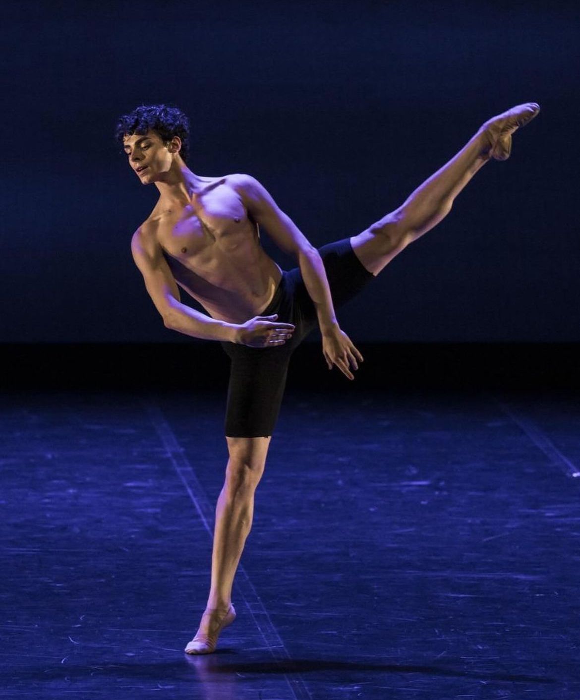 Male dancer, shirtless, in a ballet pose on a stage, leg extended high. Dark shorts. Blue lighting.