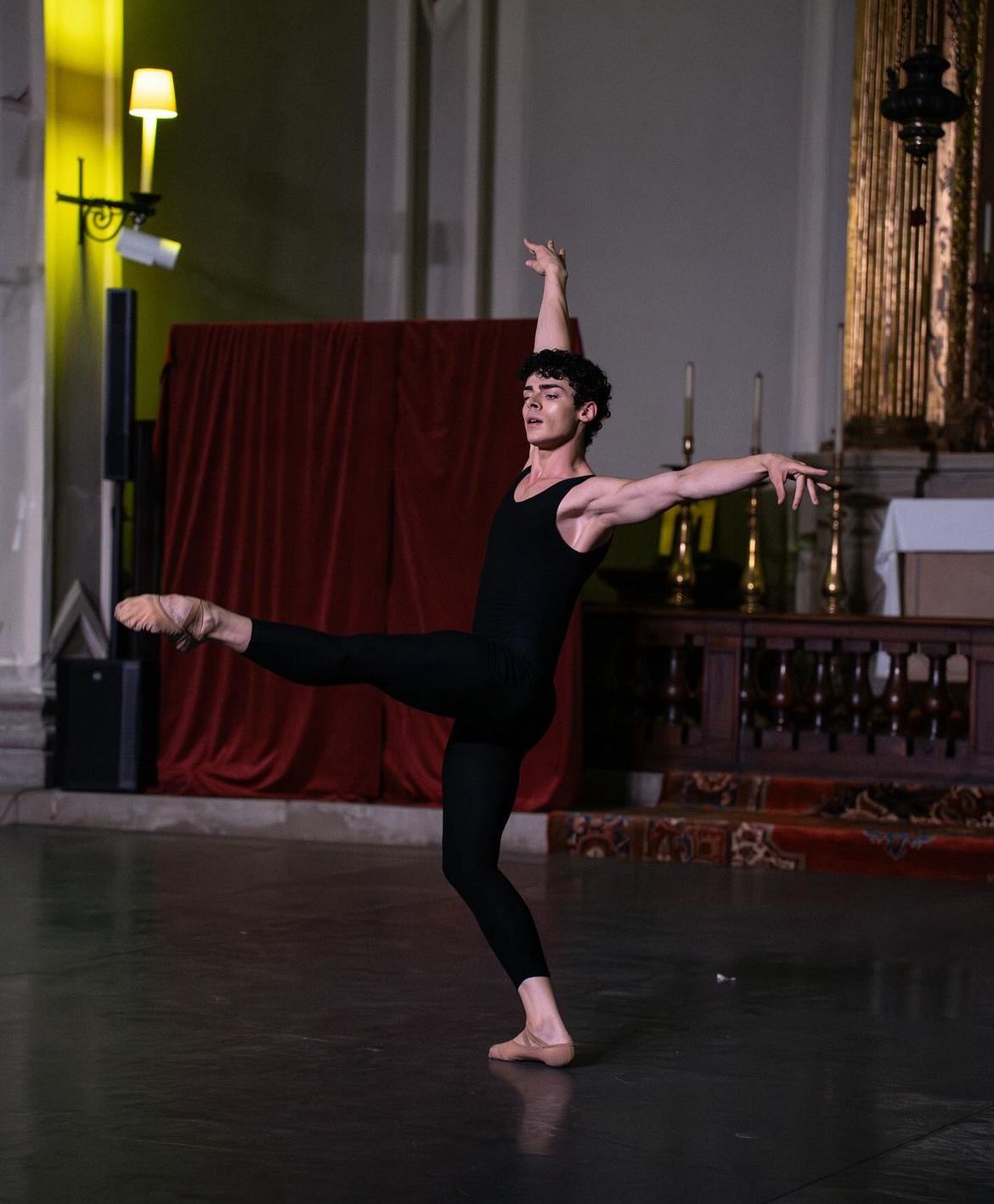 Male ballet dancer in black leotard and tights, performing a pose in a room with a red curtain.