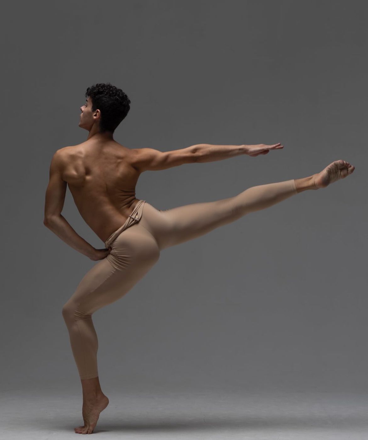 Male dancer in beige tights performing a ballet pose, arm extended, leg raised.