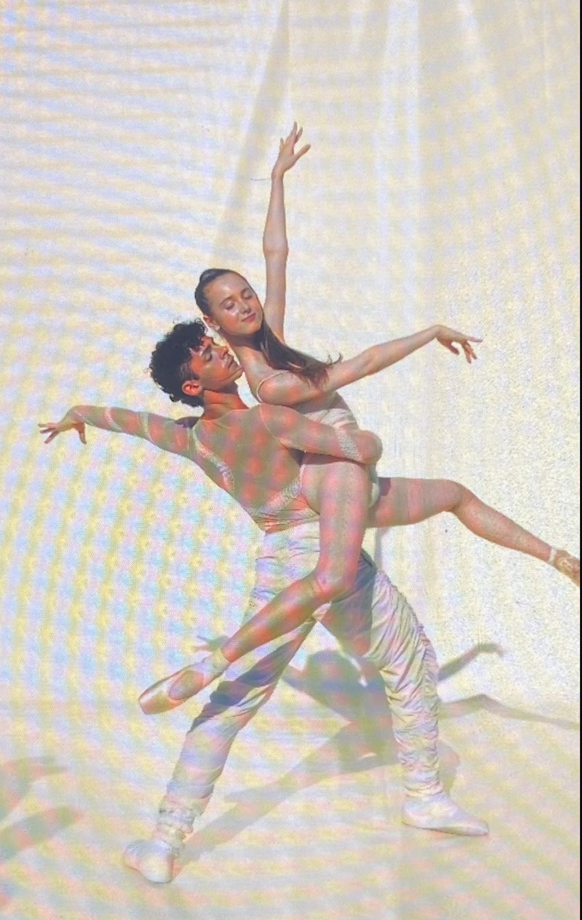 Ballet dancers in a lift pose; man supports woman. Dancers wear white, arms extended, in a light setting.