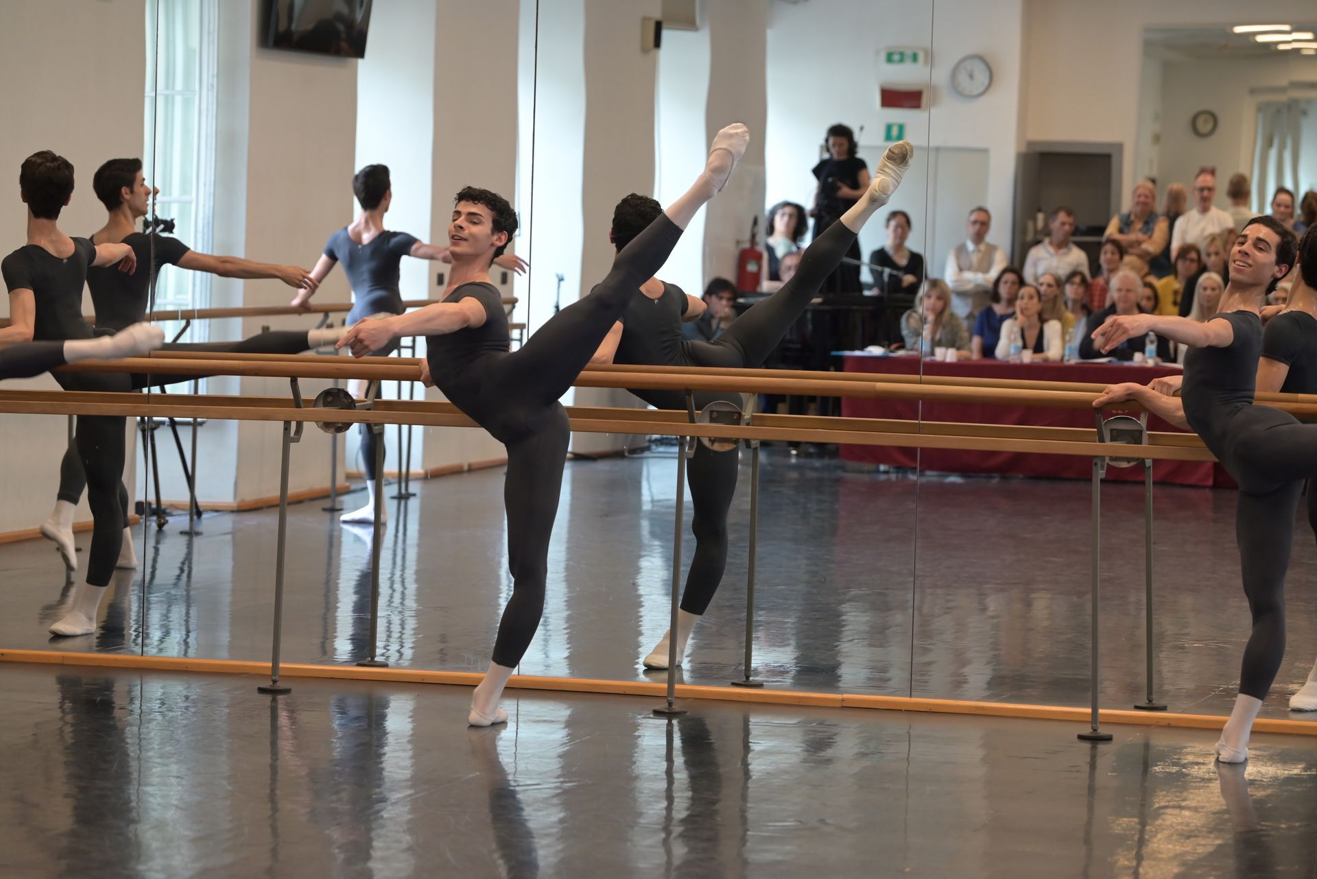 Male ballet dancers practice at a barre in a studio, leg extensions, smiles.