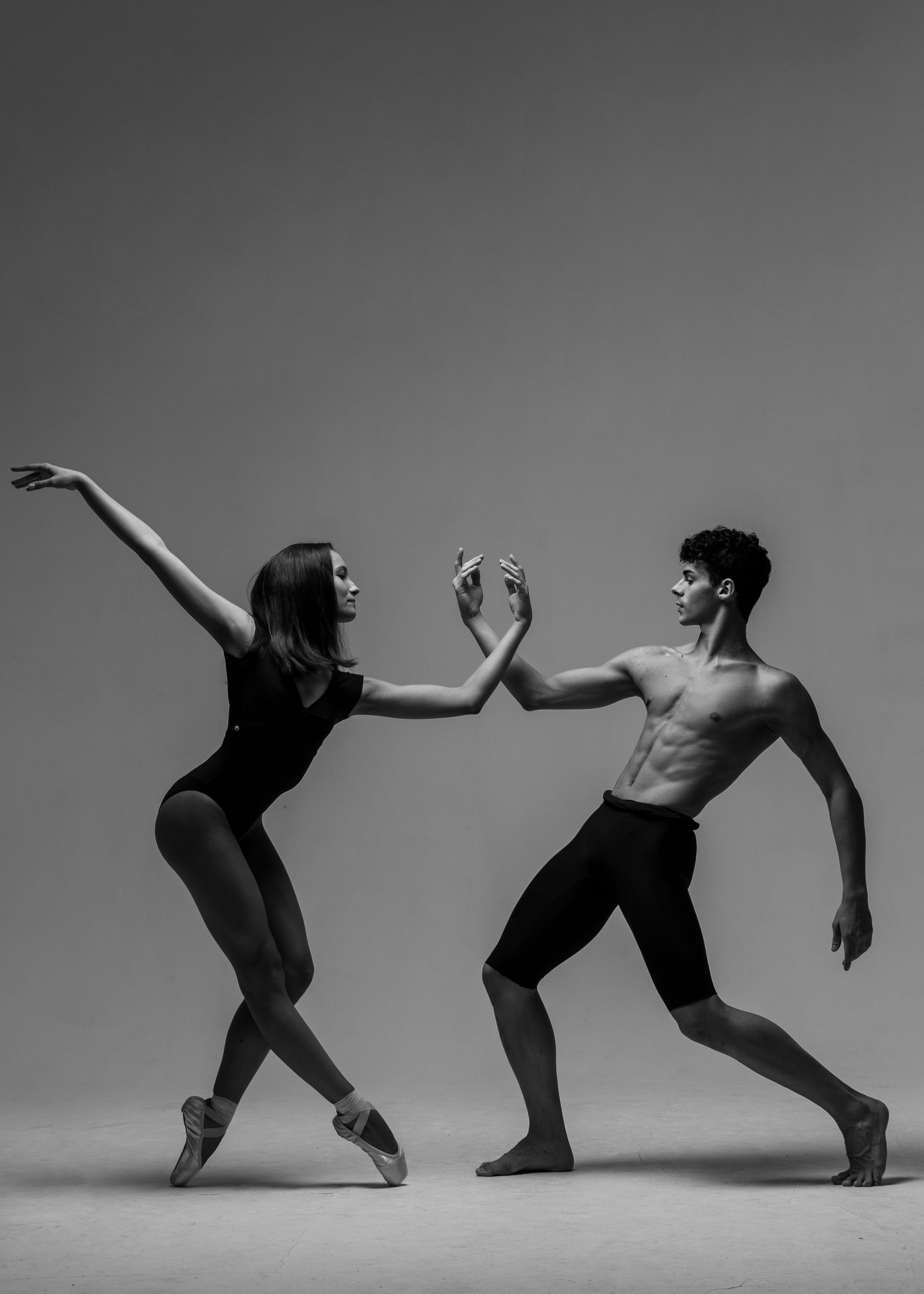 Ballet dancers in a pose; woman in leotard, man shirtless, both in motion, hands touching, gray background.