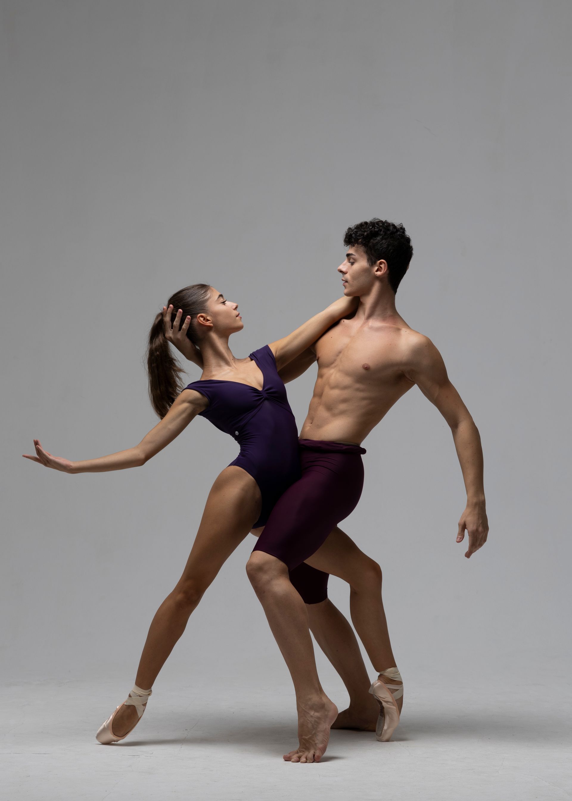Ballet dancers in a pose. Woman in purple leotard, man with bare chest, both in dance attire, gray background.