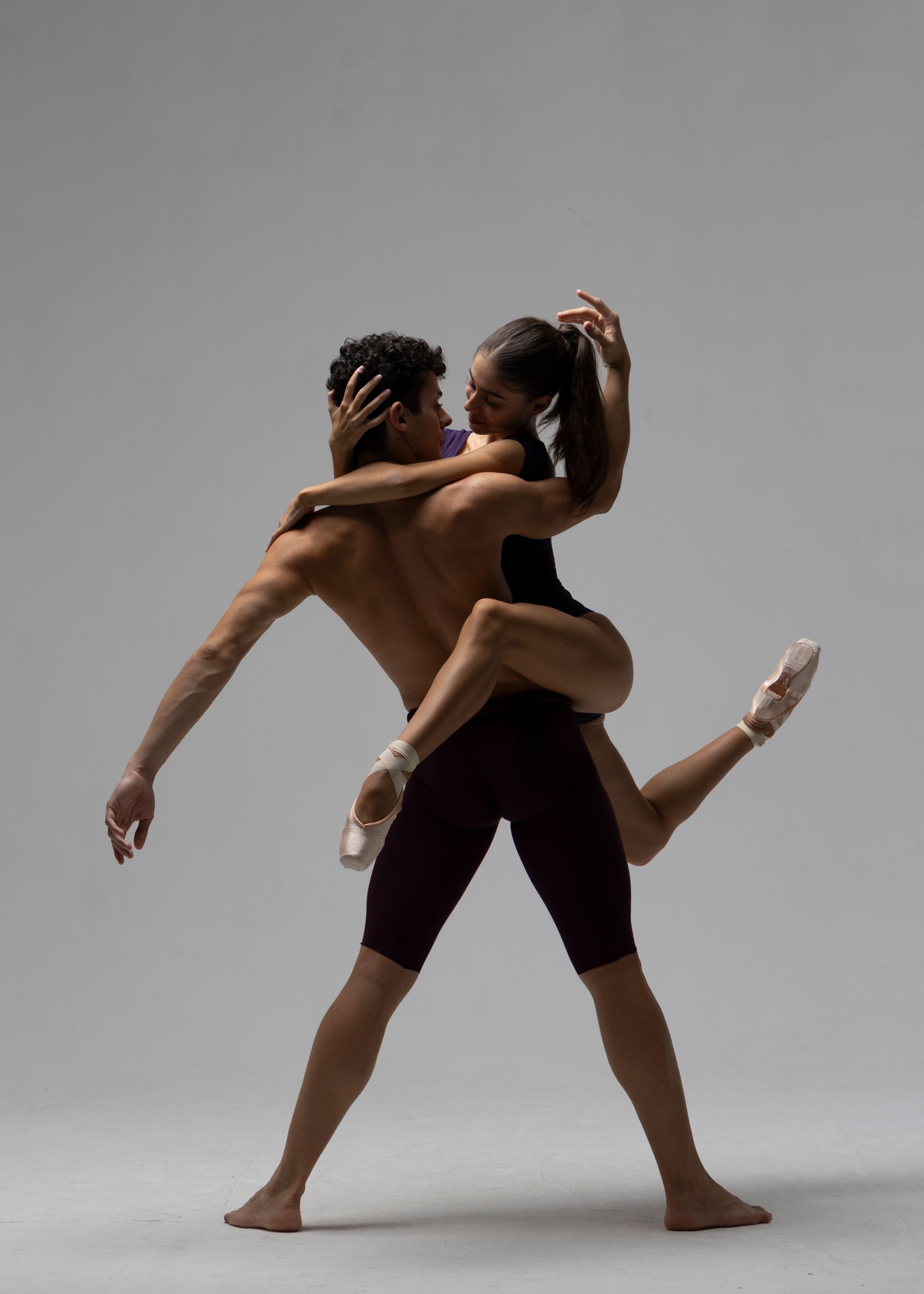 Male dancer supporting a ballerina in a backbend, wearing ballet attire, against a gray background.