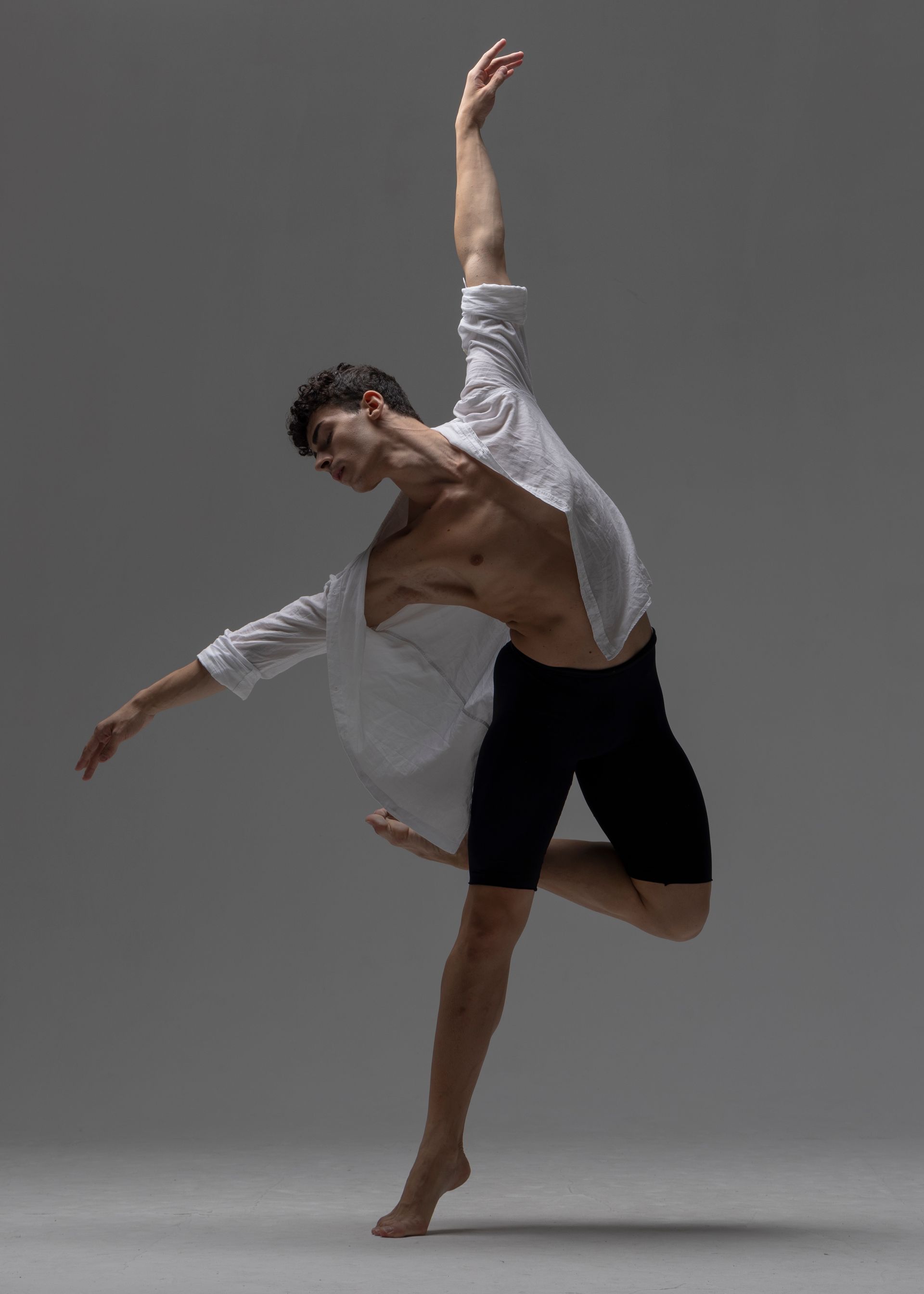 Male dancer leaping gracefully in the air, wearing a white shirt and black shorts against a gray backdrop.
