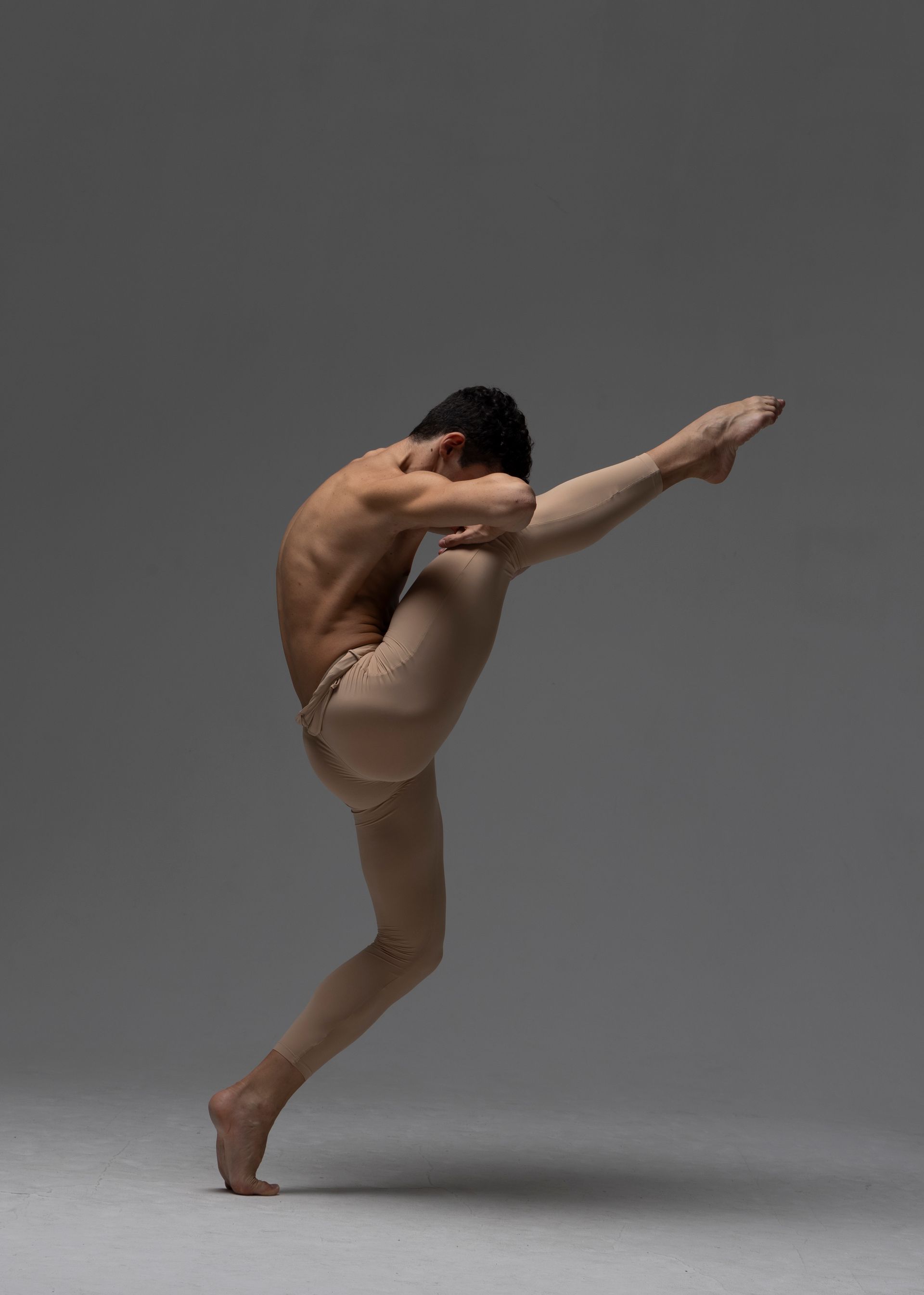 Ballet dancer in nude tights, one leg extended upward. He is bent over, arms around his other leg. Studio setting.