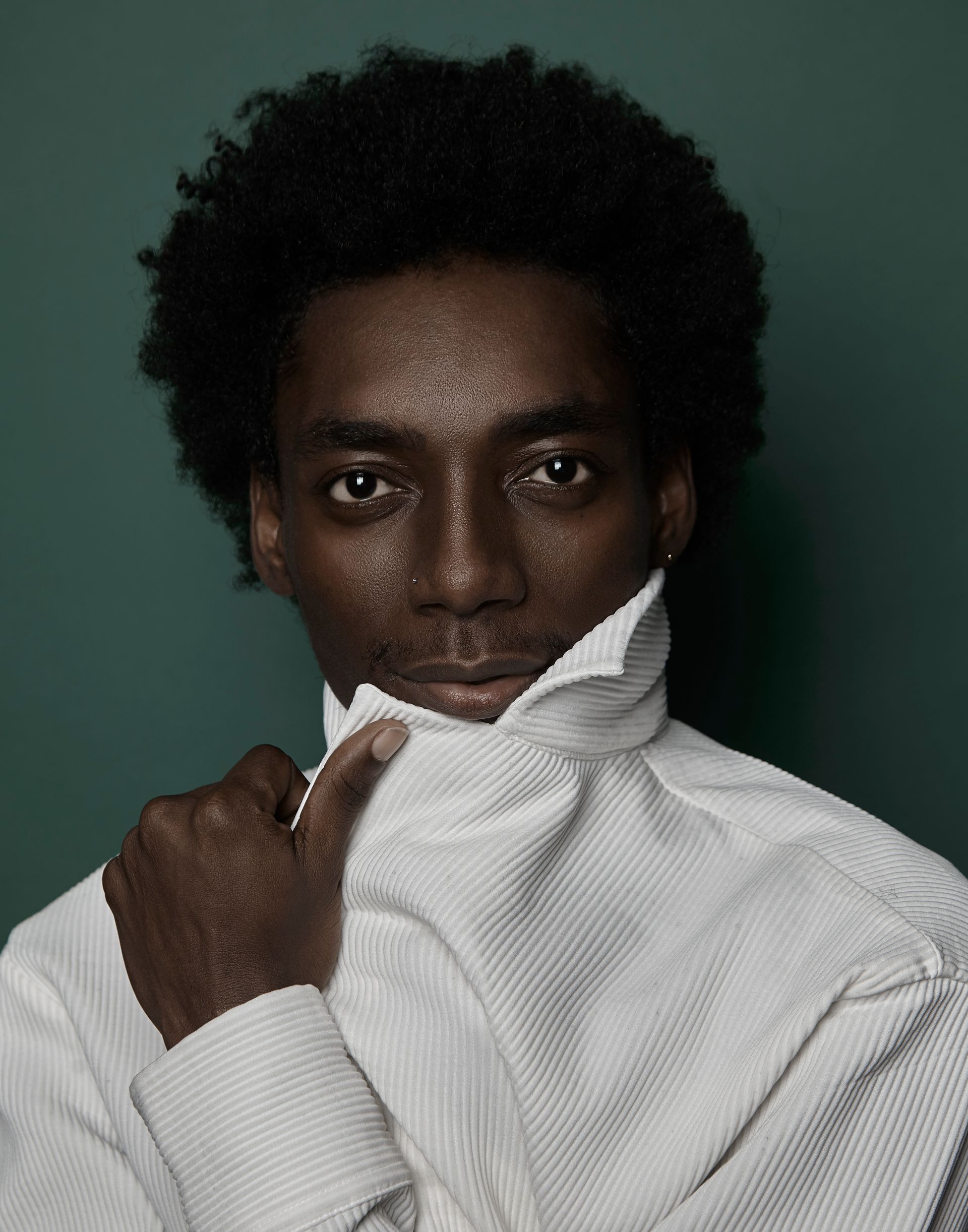 Person with dark skin, wearing a white textured shirt, pulling up the collar. Dark, curly hair. Green background.