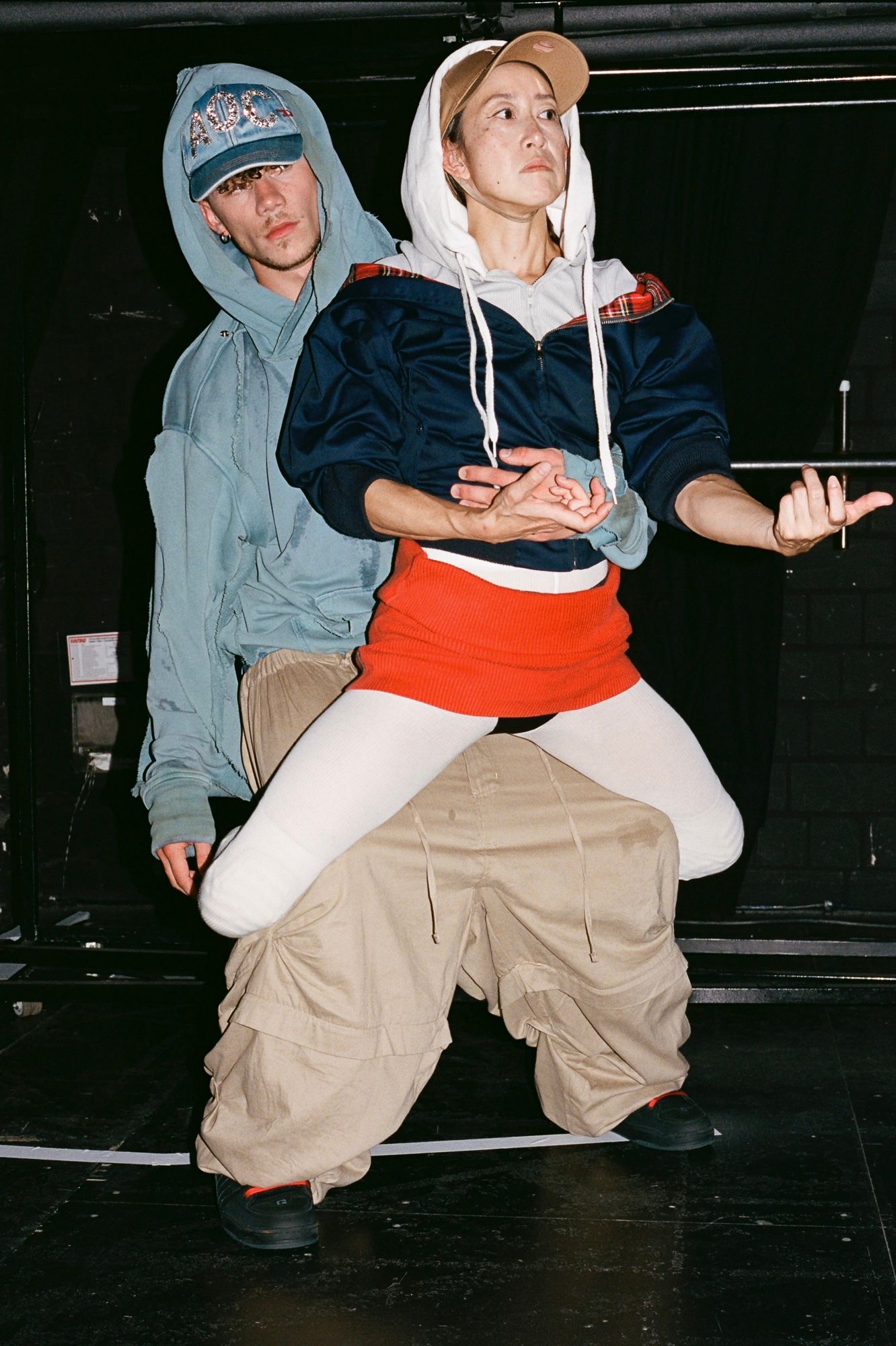 Person sitting on another's lap; both in oversized clothing, against a dark backdrop.