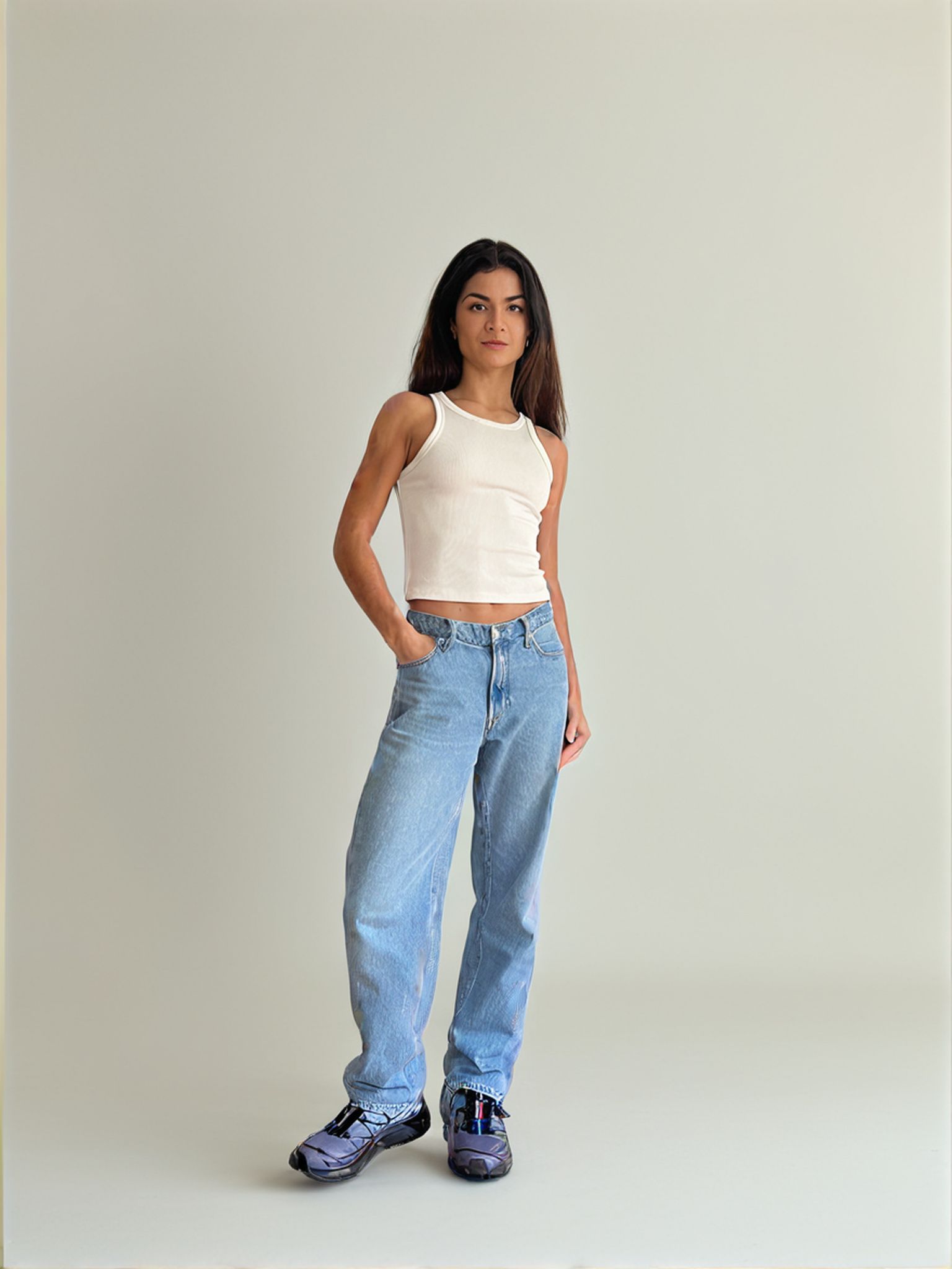 Woman in a white tank top and blue jeans, standing with one hand in her pocket, wearing sneakers.