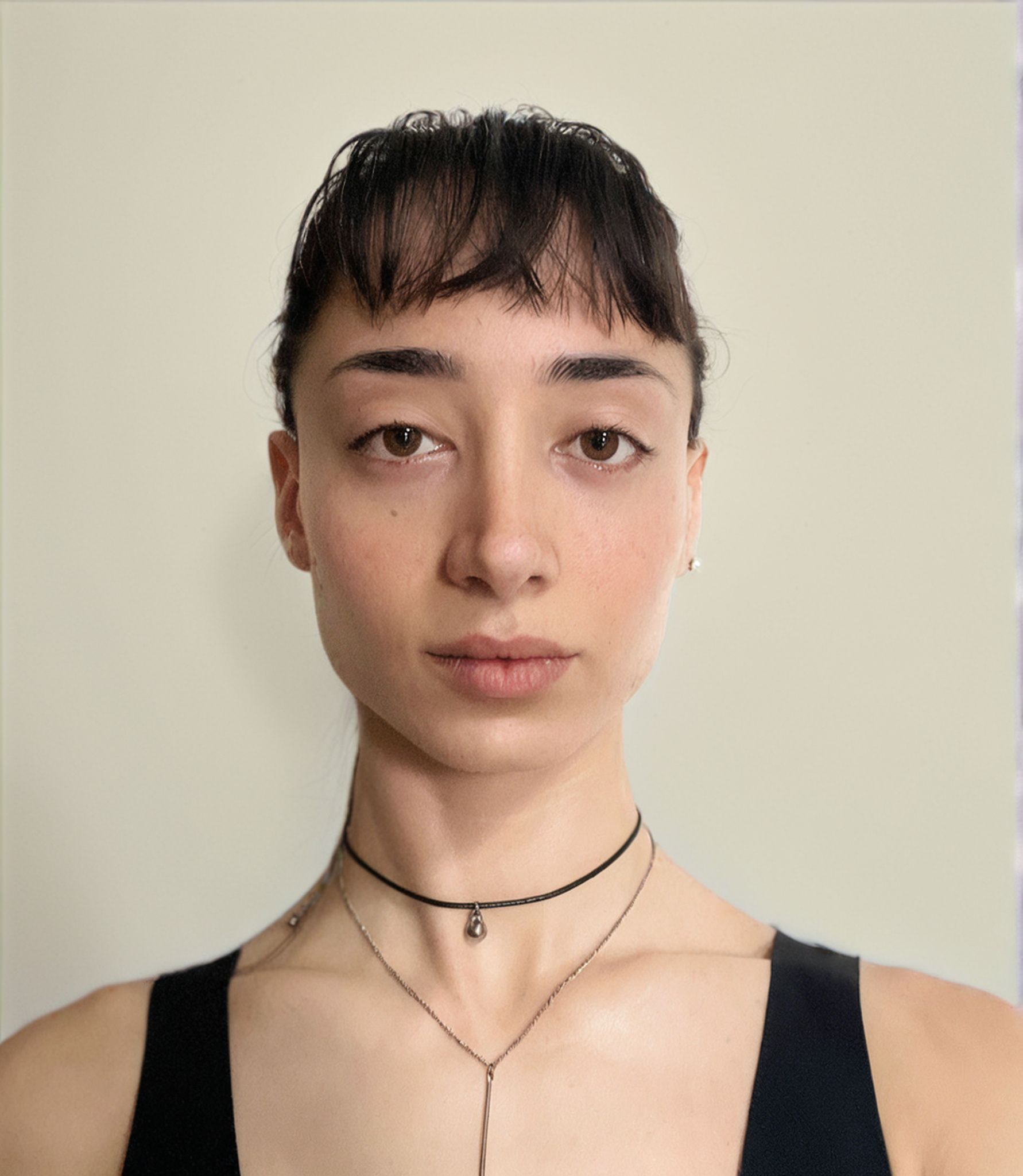 Woman with short dark hair and bangs, wearing two necklaces and a black top, against a beige background.