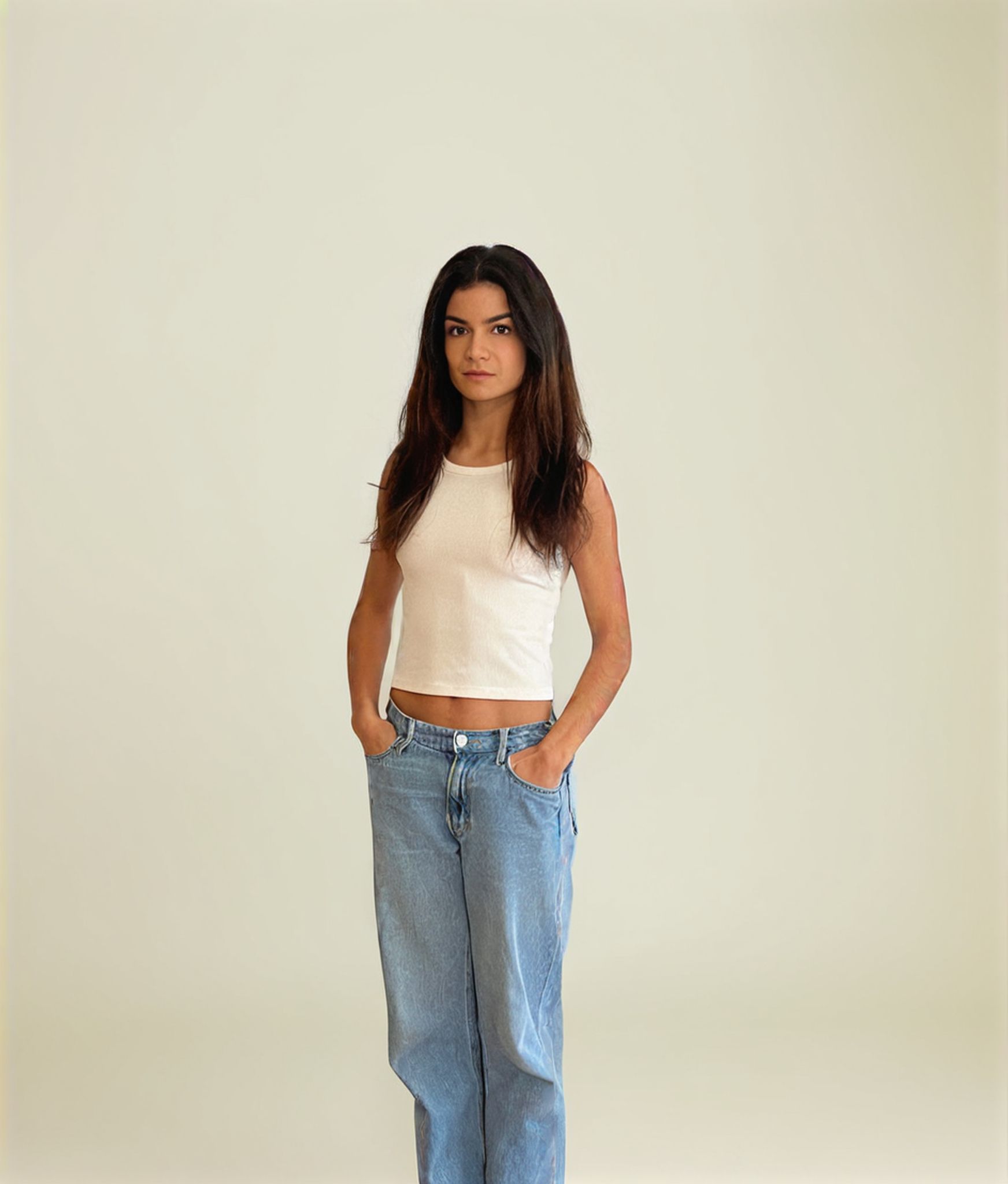 Woman in white tank top and blue jeans, hands in pockets, against a neutral background.