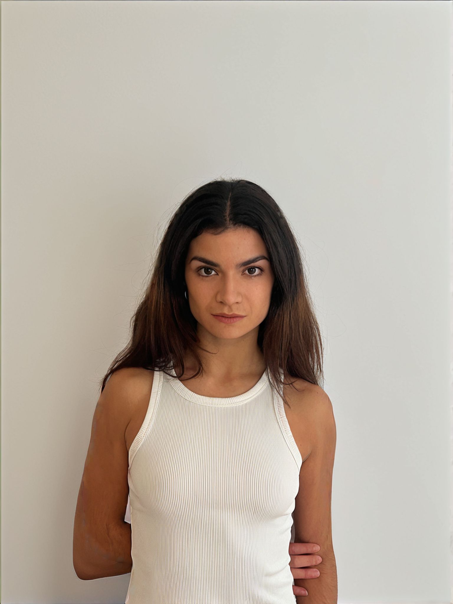 Woman in a ribbed white tank top against a white wall. Looking directly at the viewer.