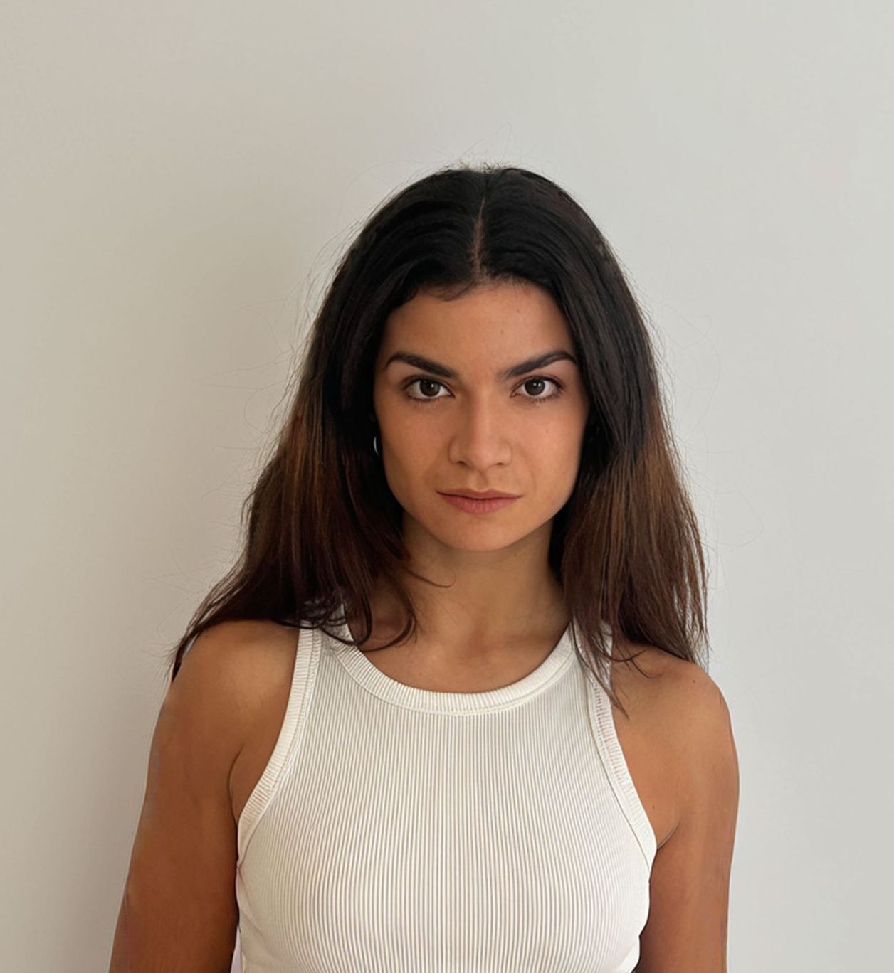 Woman with dark hair in a white ribbed tank top, looking directly at the camera against a white wall.