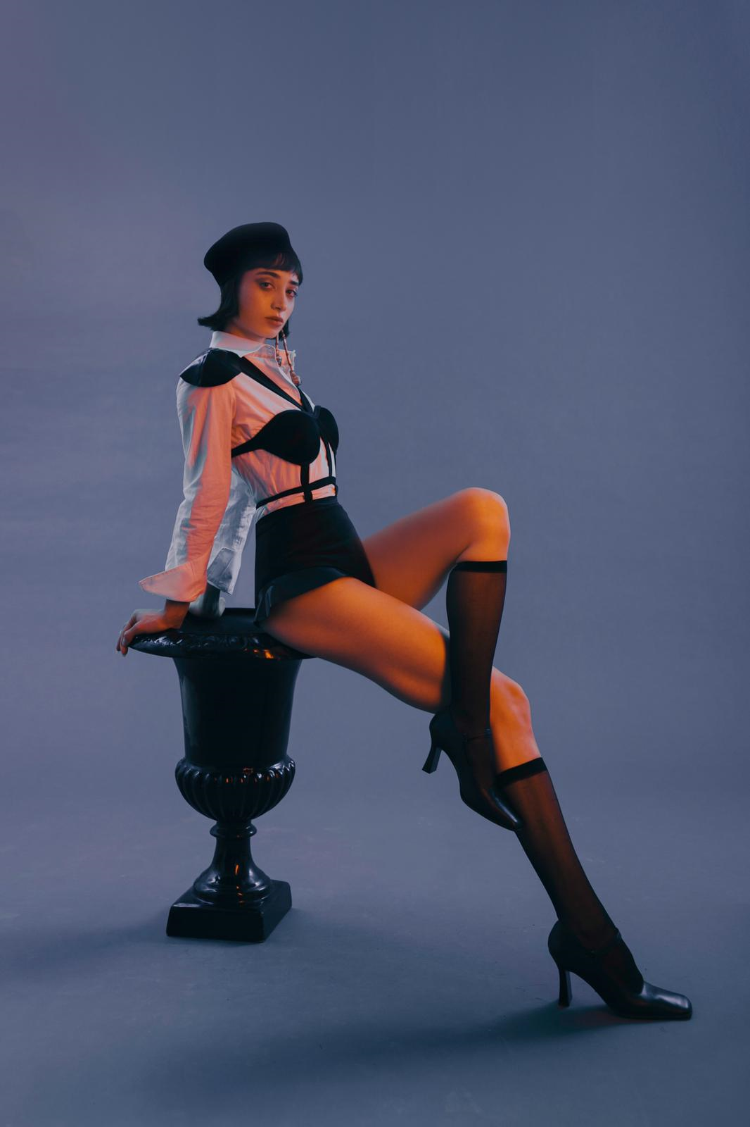 Woman in black and white outfit poses on ornate black vase against blue background.