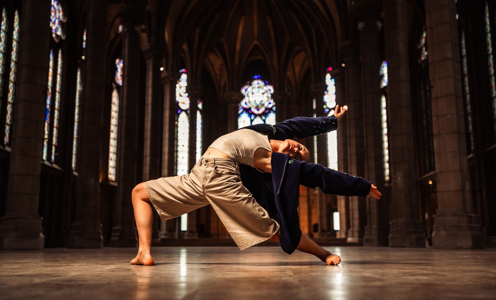 Dancer in beige and navy arched backwards, arms outstretched, inside a cathedral with stained-glass windows.