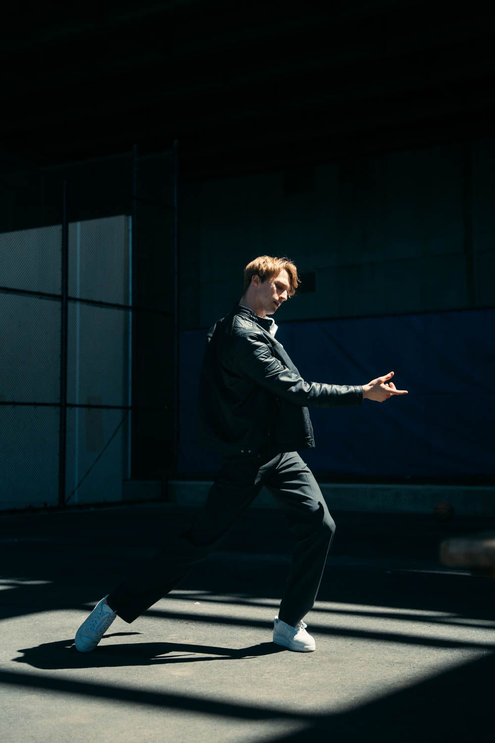 Man in black jacket and pants, dancing, angled view, outdoor setting, sunlight.