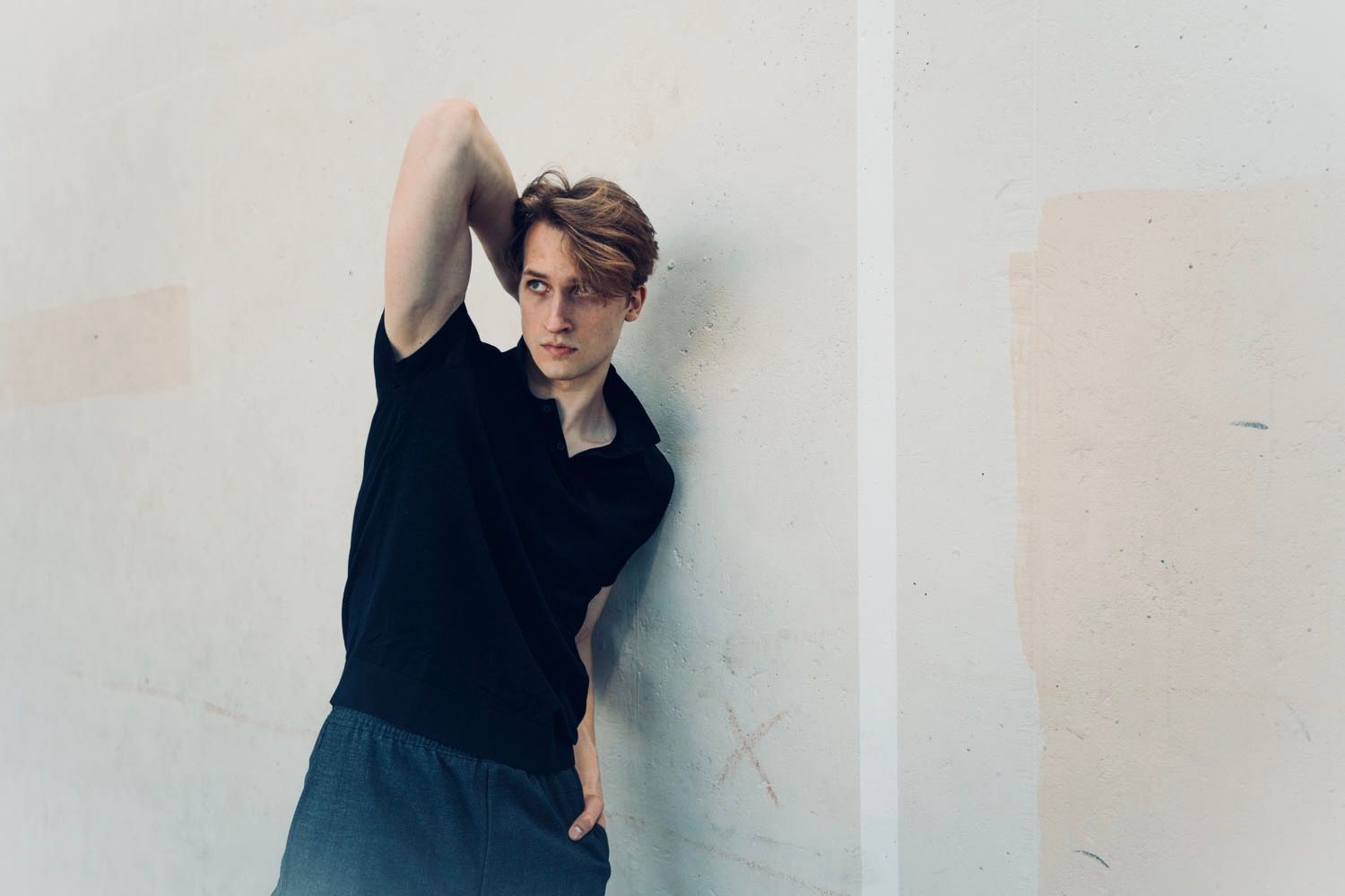 Man in black shirt and blue pants, leaning against a white wall, arm raised.
