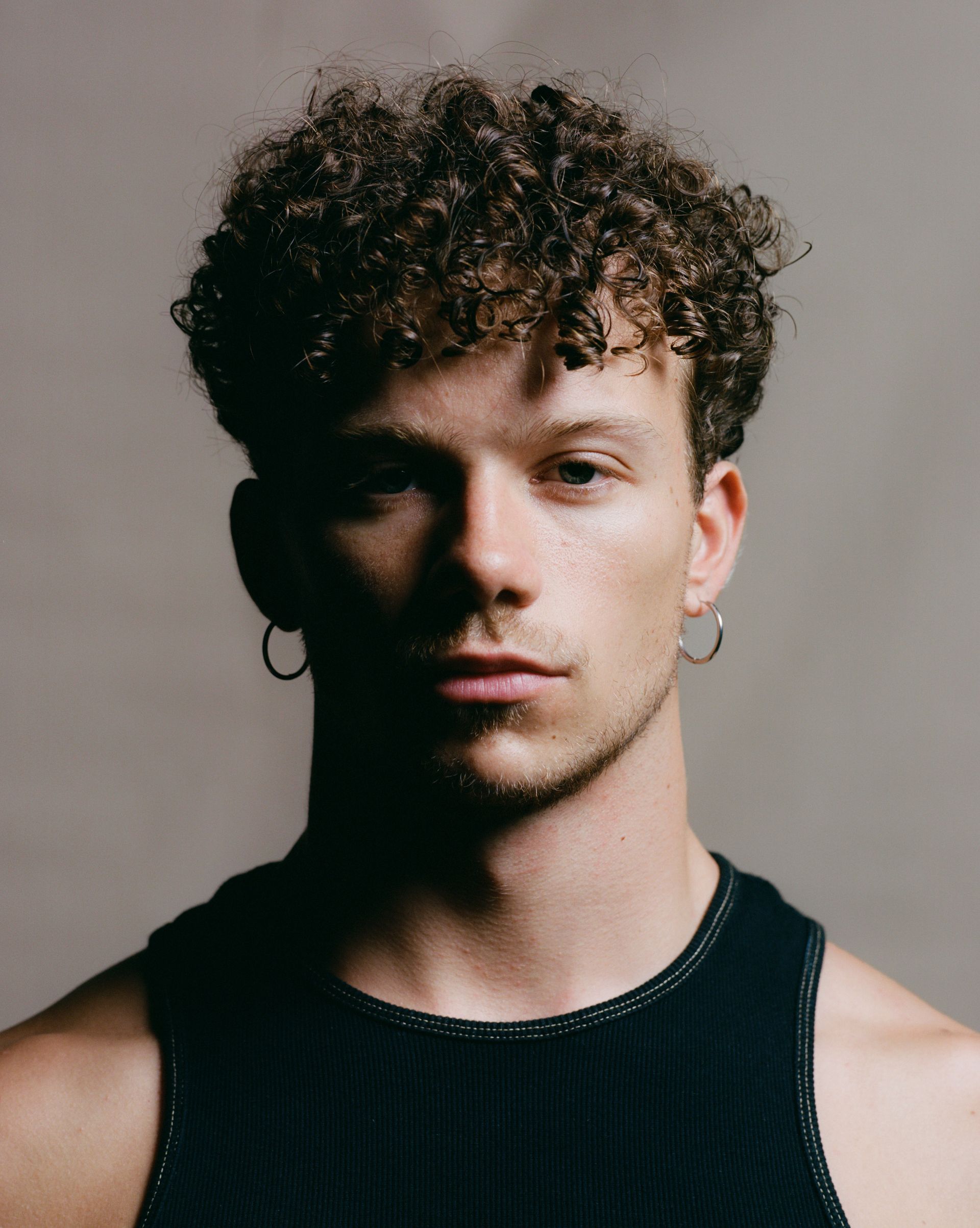 Man with curly hair and hoop earrings, wearing a black tank top, looking directly at the camera.