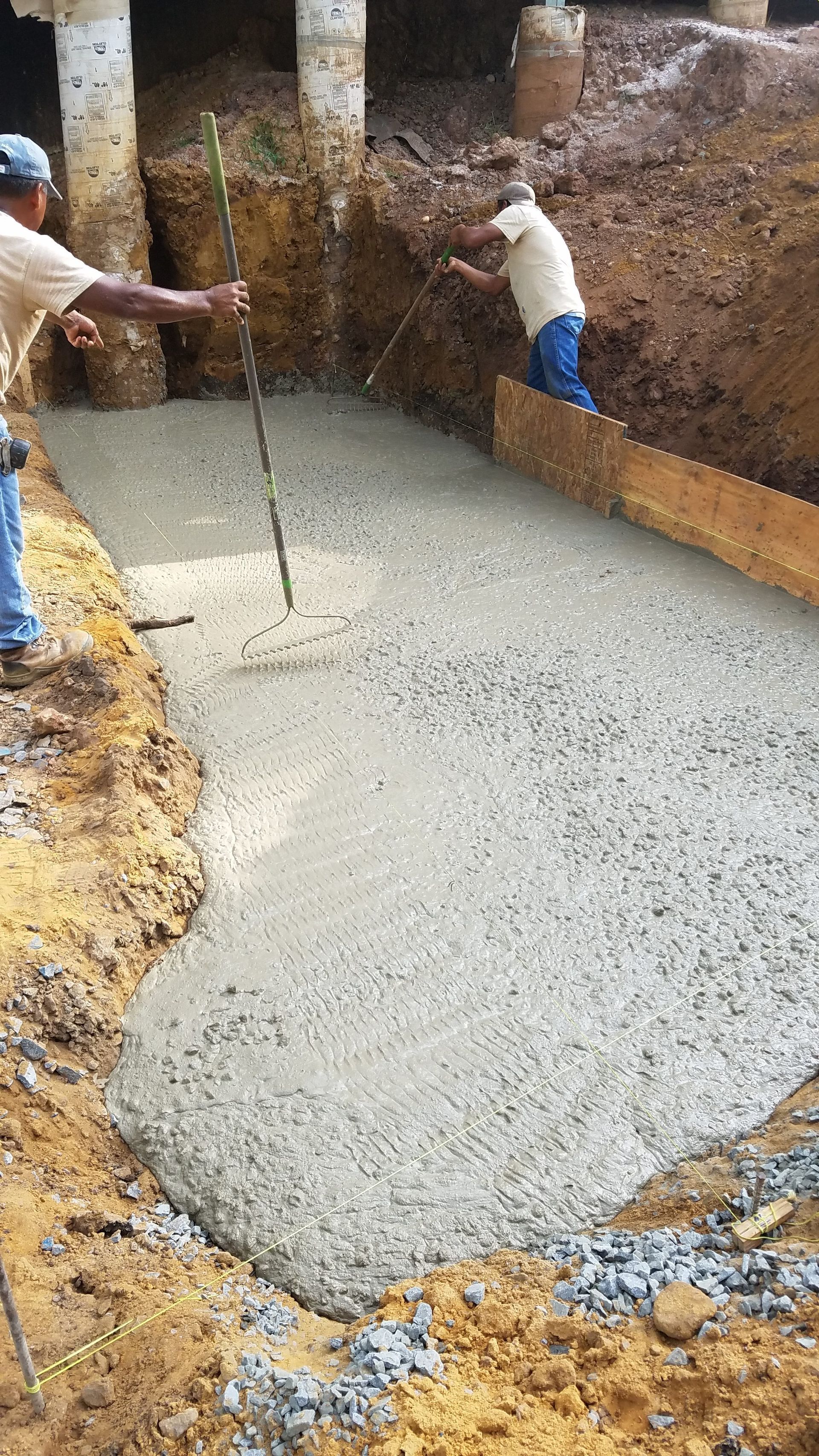 Construction workers pouring concrete foundation, dirt excavation, sunny day.