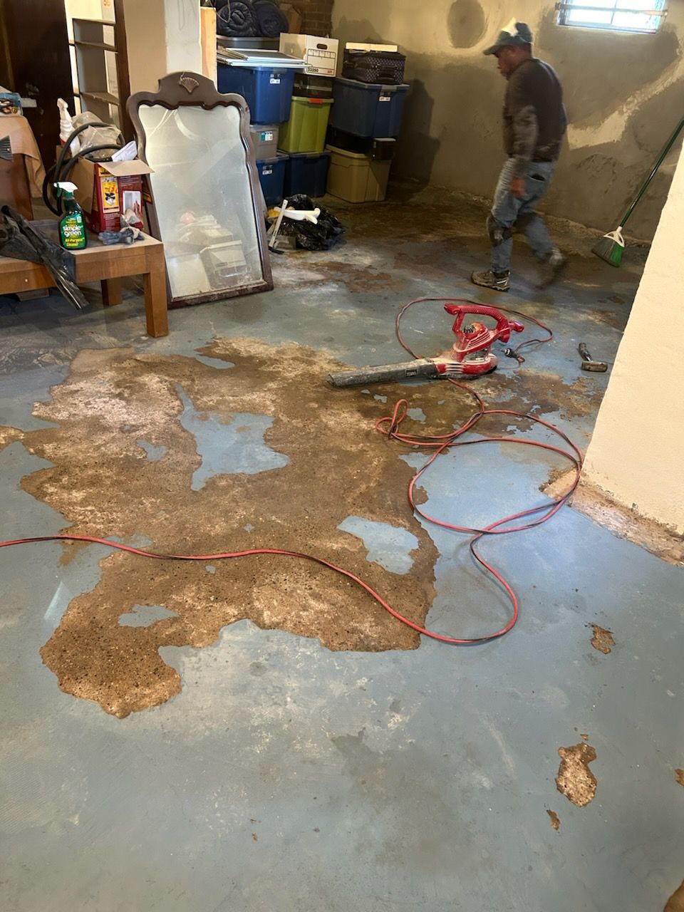 A man in a basement using a leaf blower on a large spill of wet brown material.