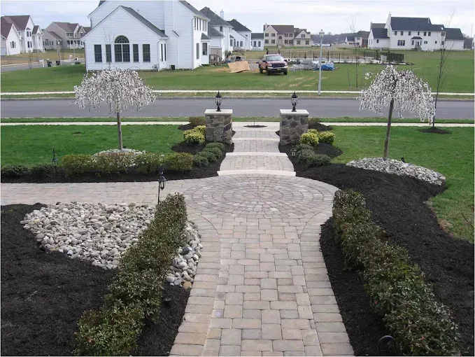 Brick pathway leading to a home, with landscaped gardens, trees, and houses in the background.