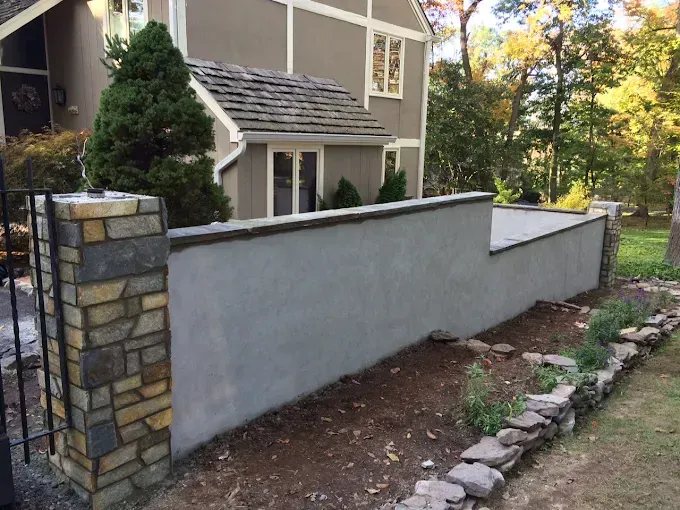 Stone pillars frame a gray stucco wall with a dark trim; house in the background.