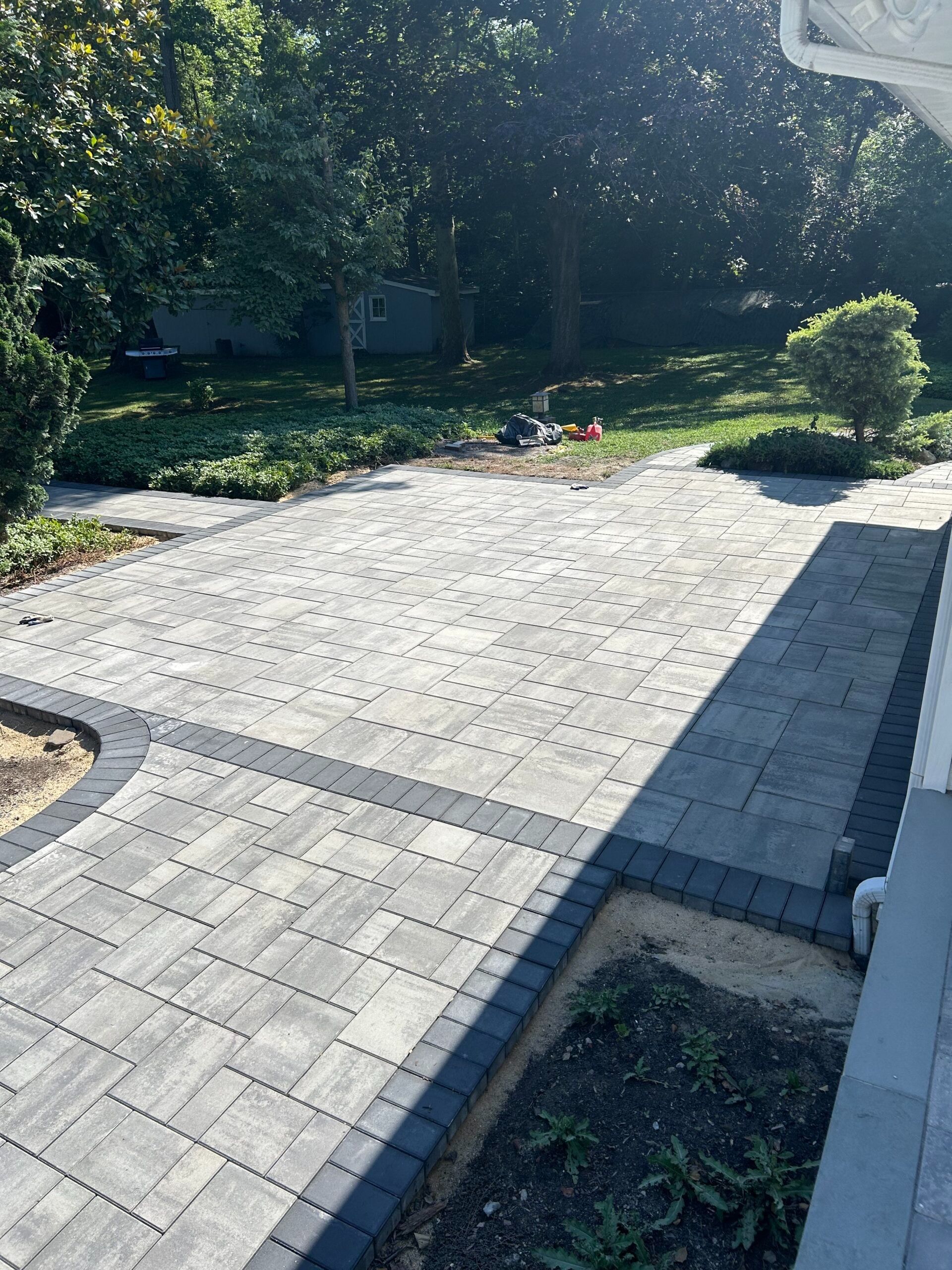Paved patio and walkway with contrasting dark border, leading to a grassy yard with trees.