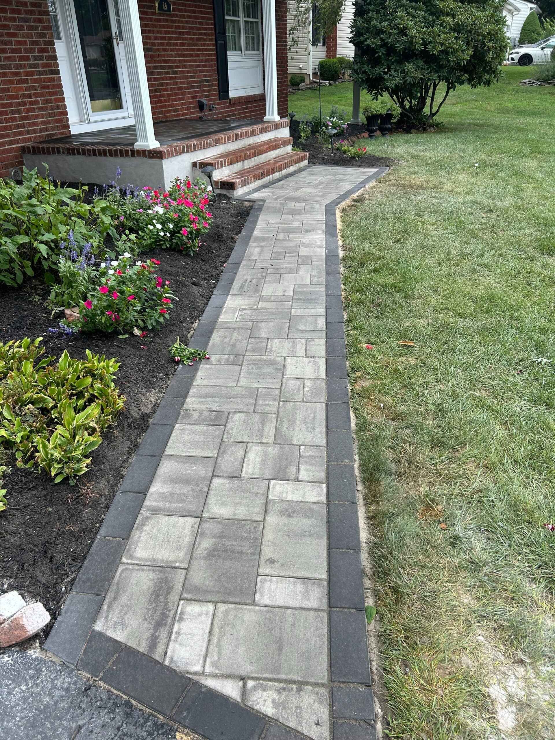 Brick walkway leading to a house entrance with a flower bed on one side and grass on the other.