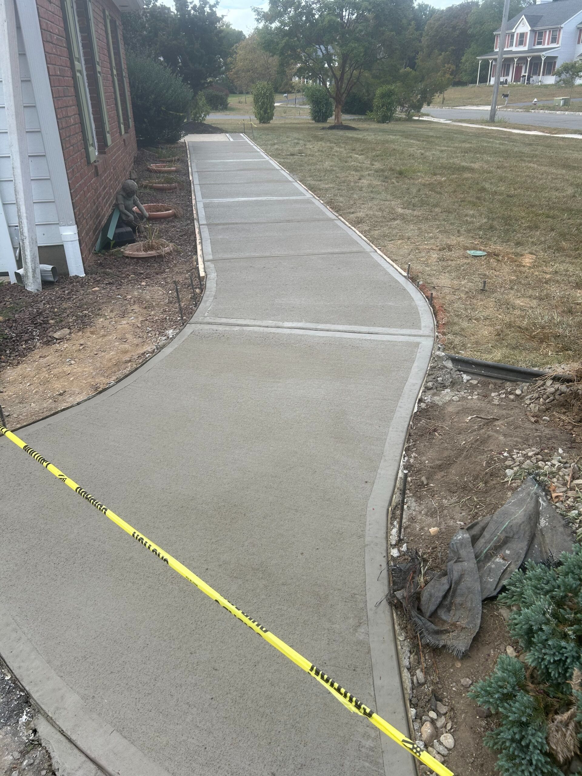 New concrete sidewalk curving along a house; yellow caution tape in the foreground.