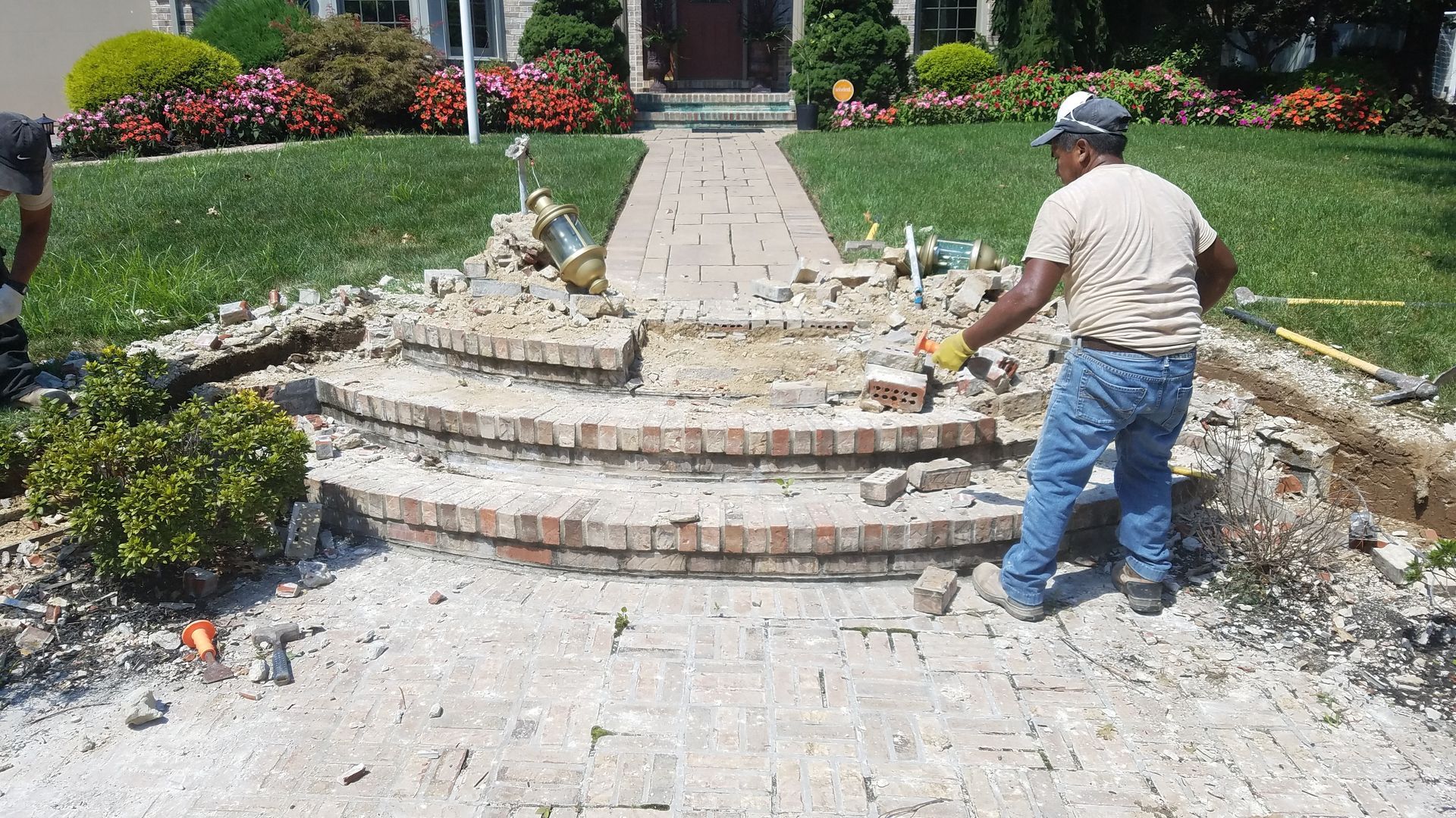 Workers demolishing brick steps and walkway in front of a house, on a sunny day.
