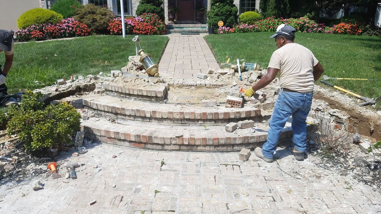 Workers demolishing brick steps and walkway in front of a house, on a sunny day.