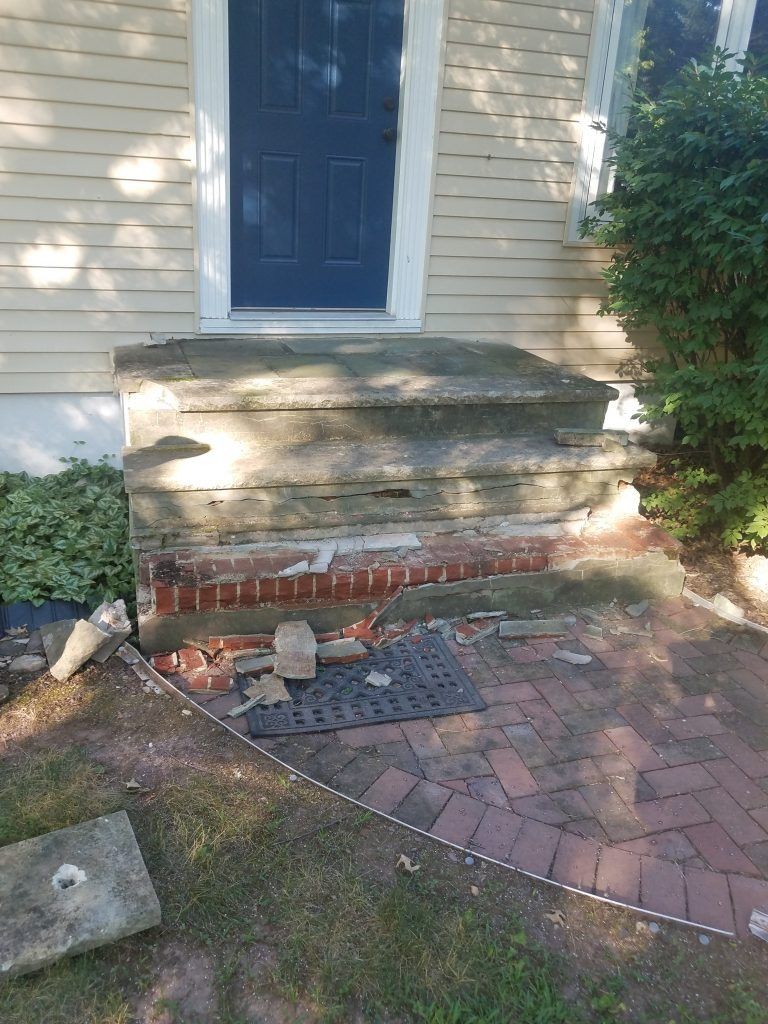 Damaged front porch with crumbling concrete and exposed brick, leading to a blue door.