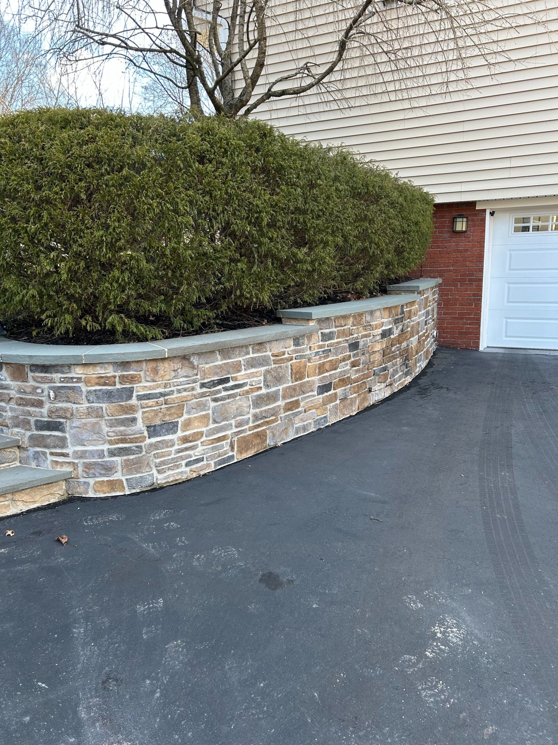 Stone retaining wall with green bushes, beside a driveway and garage.