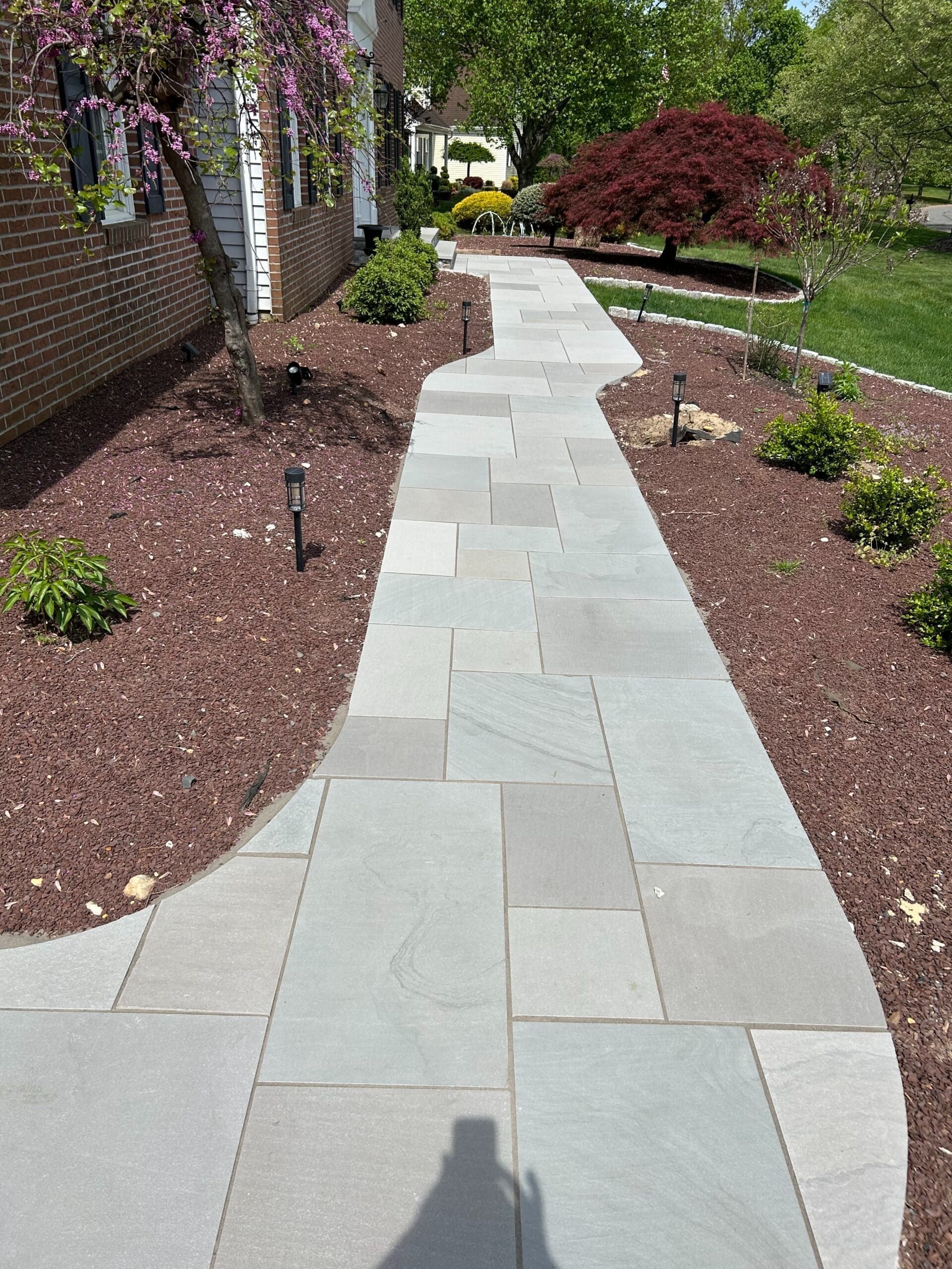 Stone pathway leading to a house, bordered by brown mulch and green bushes; red tree in the background.