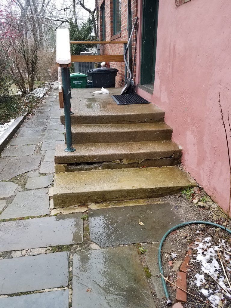 Concrete steps leading to a doorway, with a handrail and wet stone walkway.