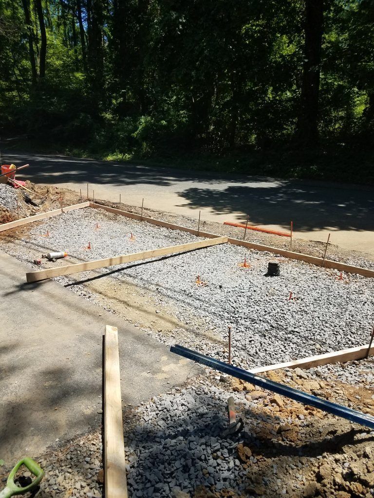 Construction site with gravel-filled formwork for a concrete slab, next to a paved road in a wooded area.
