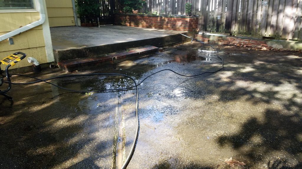 Wet concrete patio with black hose, partial view of a yellow house and a brick planter.